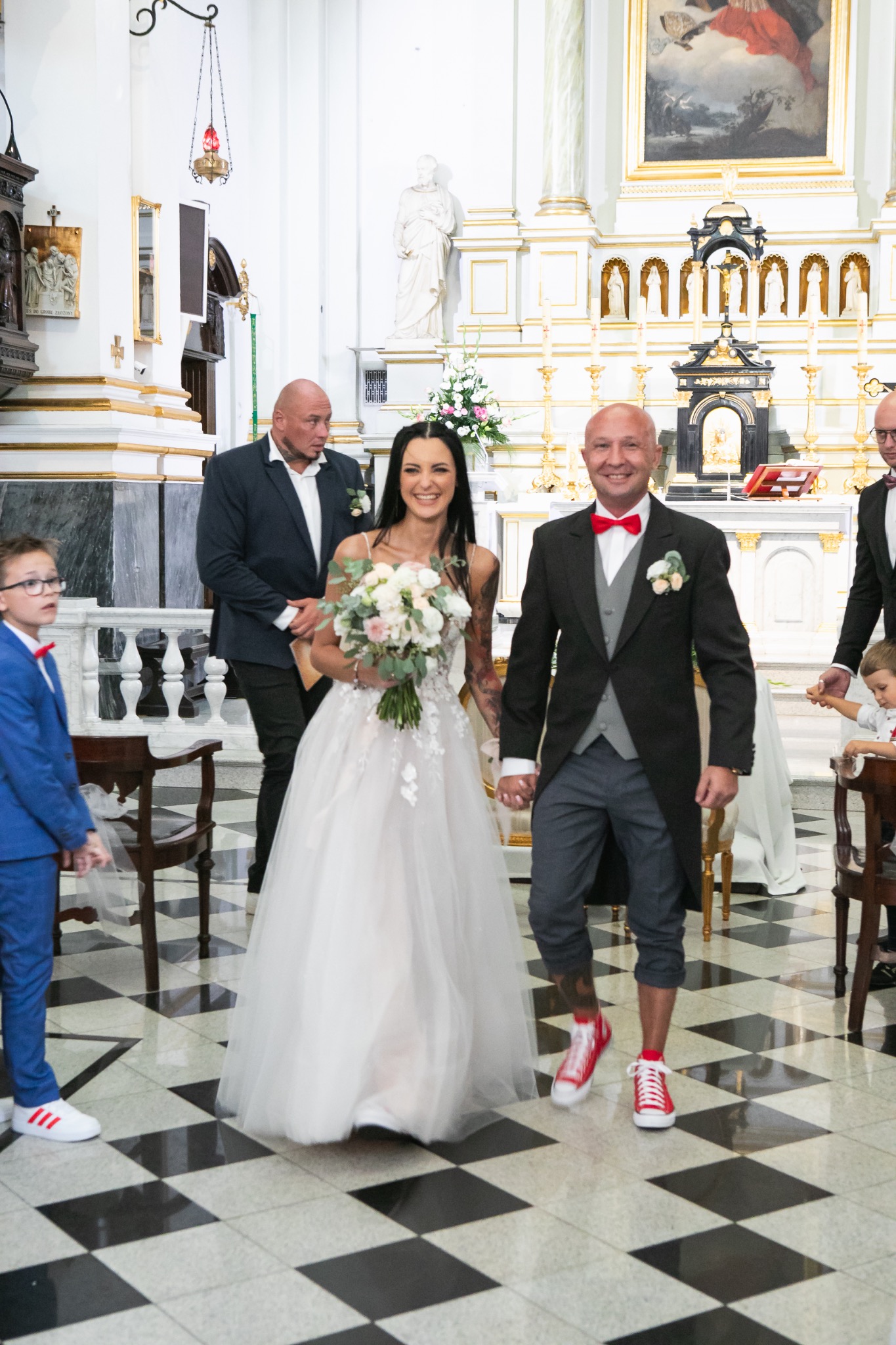 Uśmiechnięta para młoda, trzymająca się za ręce, w kościele. Pan młody w sportowych butach. W tle goście i czarno-biała podłoga.