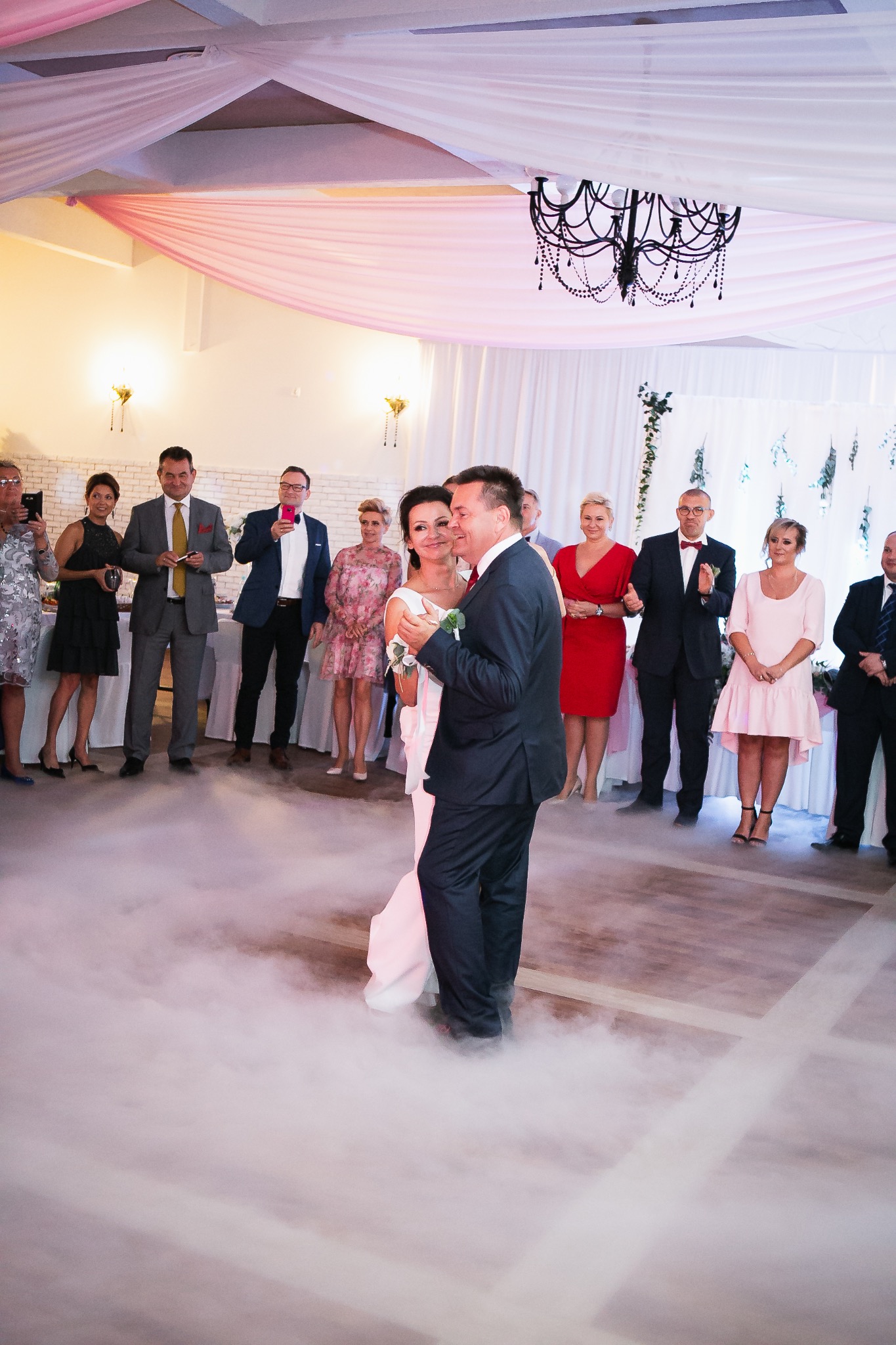 Para młoda tańczy pierwszy taniec w chmurach dymu, otoczona gośćmi weselnymi w eleganckiej sali z dekoracjami i żyrandolem.