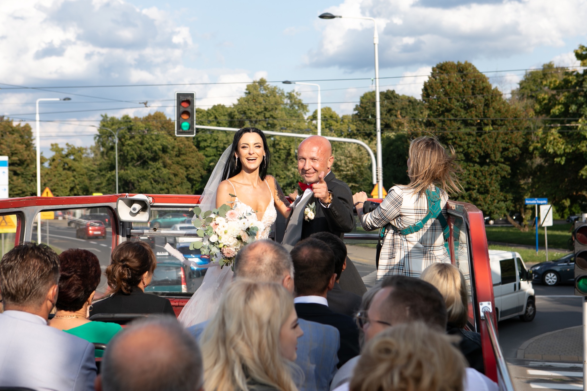 Uśmiechnięta para młoda w otwartym autobusie z gośćmi, on w czarnym garniturze z muszką, ona w białej sukni z bukietem, w tle sygnalizacja świetlna i zieleń.
