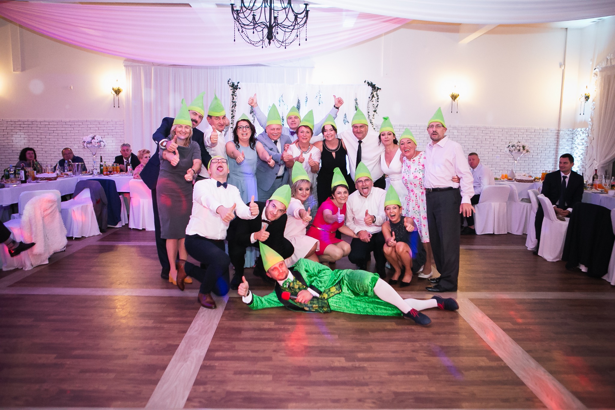Grupa osób w zielonych czapkach pozuje do zdjęcia w sali bankietowej z parkietem. Jeden mężczyzna leży na podłodze w zielonym przebraniu.