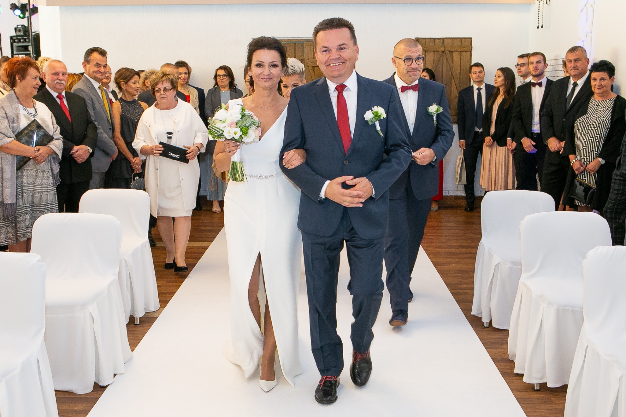 Uśmiechnięta para młoda prowadzona do ołtarza przez świadków, w tle goście weselni. Biały dywan i krzesła w pokrowcach. Zdjęcie z uroczystości ślubnej.