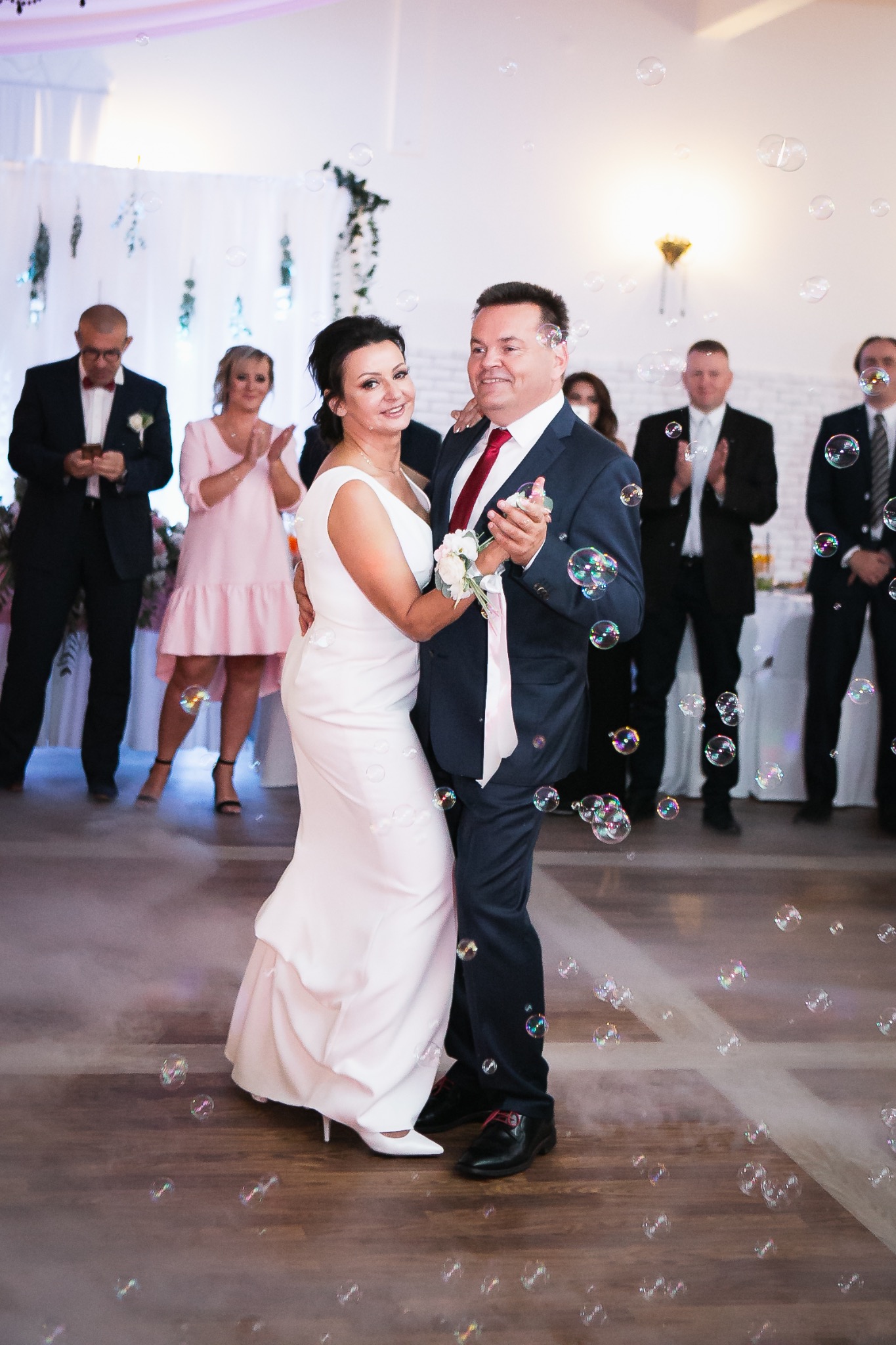 Para młoda w tańcu, otoczona bańkami mydlanymi, goście w tle. Ujęcie z wesela, sukienka panny młodej i garnitur pana młodego, dekoracje sali.