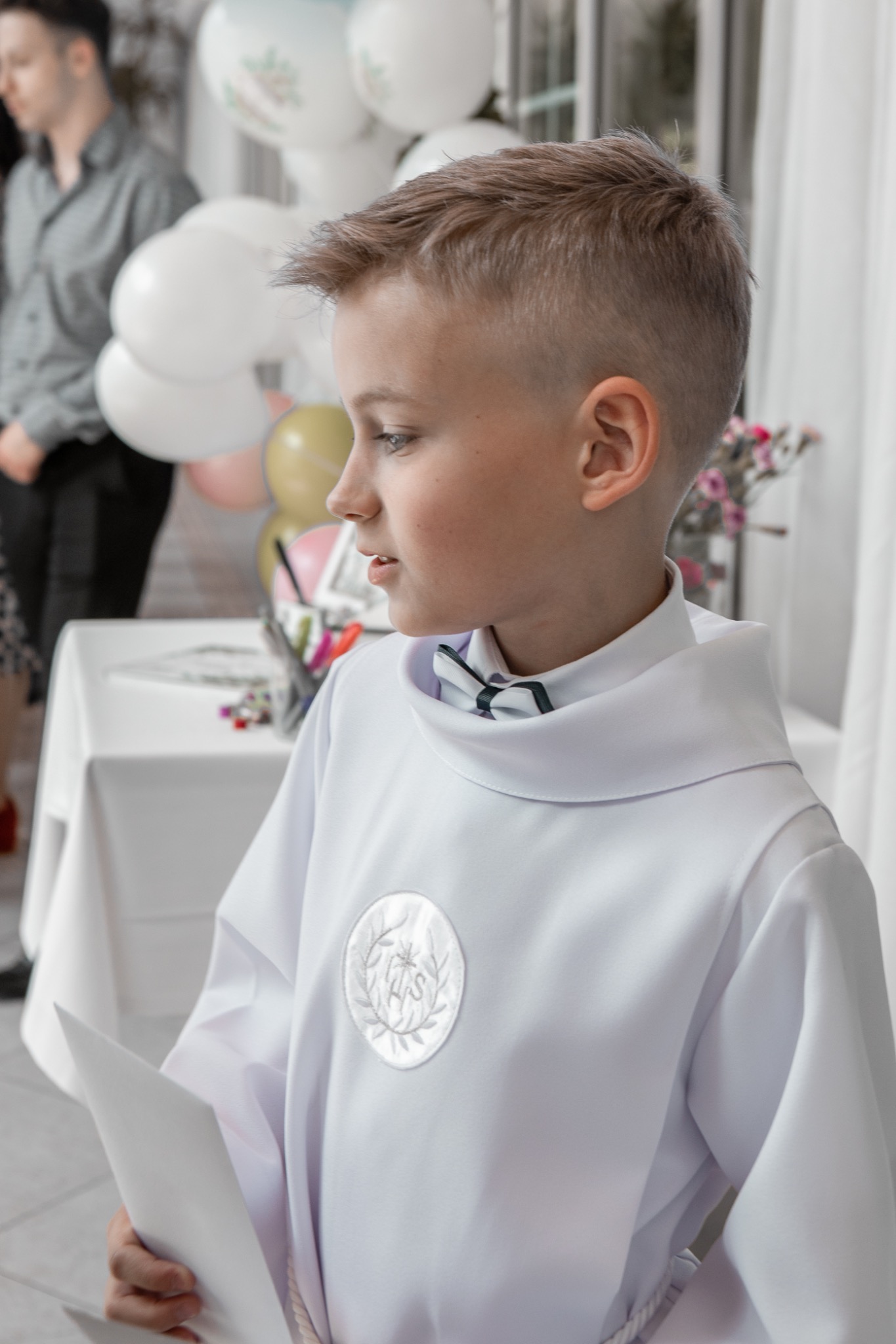 Portret chłopca w stroju komunijnym z muchą, trzymającego kartkę, w jasnym wnętrzu z dekoracjami. Ujęcie z profilu, skupiające się na detalu stroju i wyrazie twarzy.