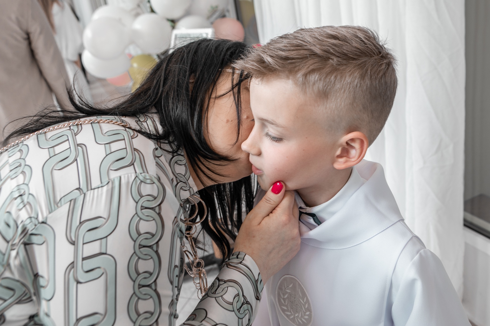 Matka całuje syna ubranego w białą albę komunijną. W tle białe balony i zasłony. Ujęcie z bliska, skupione na emocjach.