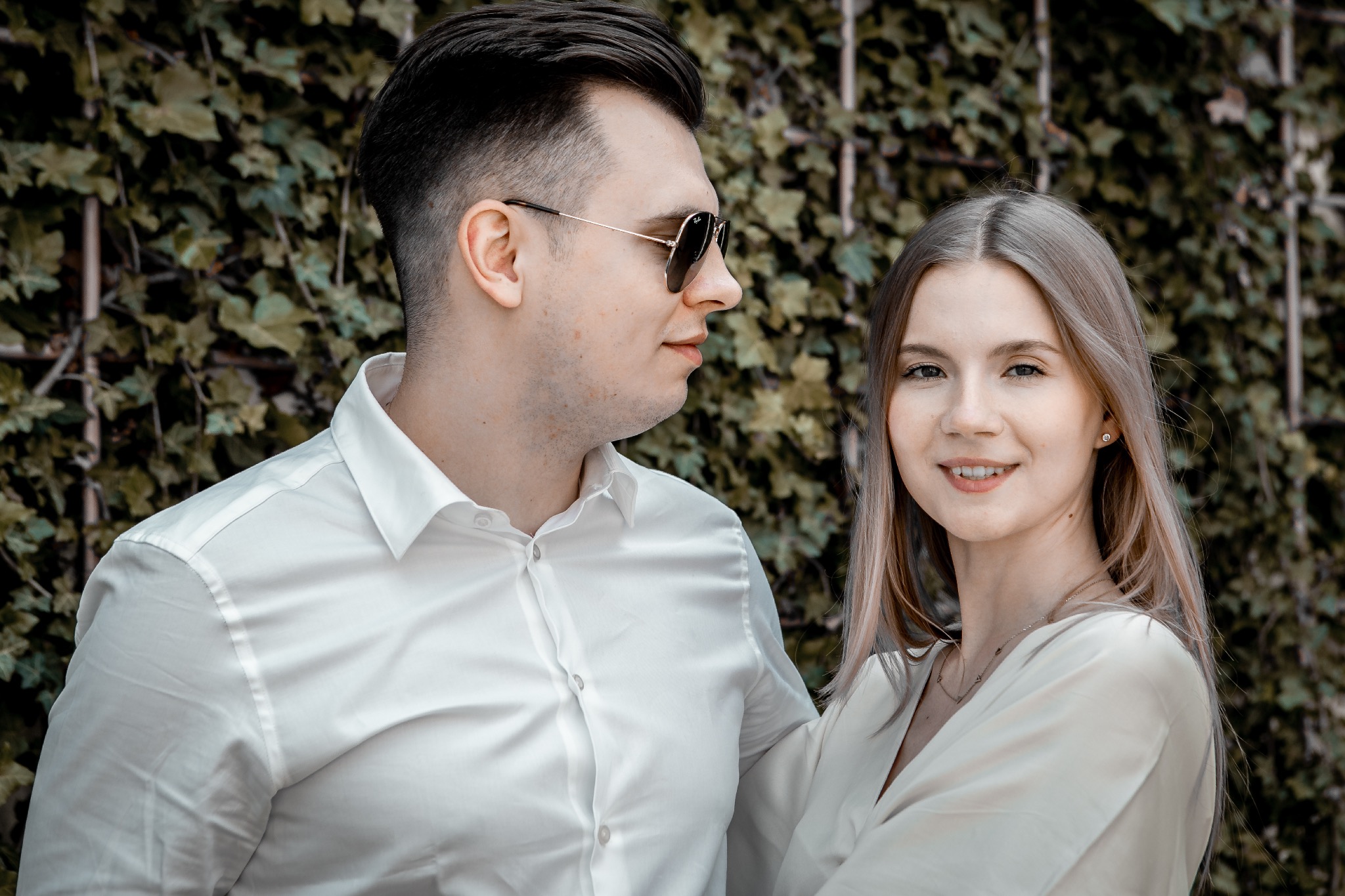 Para w białych koszulach pozuje na sesji zdjęciowej na tle ściany z bluszczu. Mężczyzna w okularach spogląda w bok, kobieta uśmiecha się do kamery.
