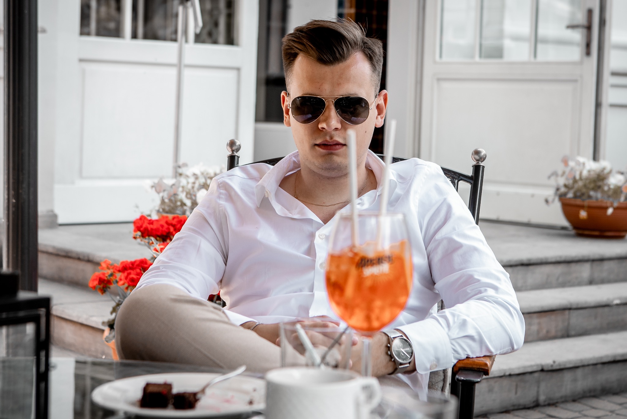 Mężczyzna w okularach przeciwsłonecznych siedzi przy stoliku z drinkiem Aperol Spritz, deserem i kawą. W tle białe drzwi i kwiaty.