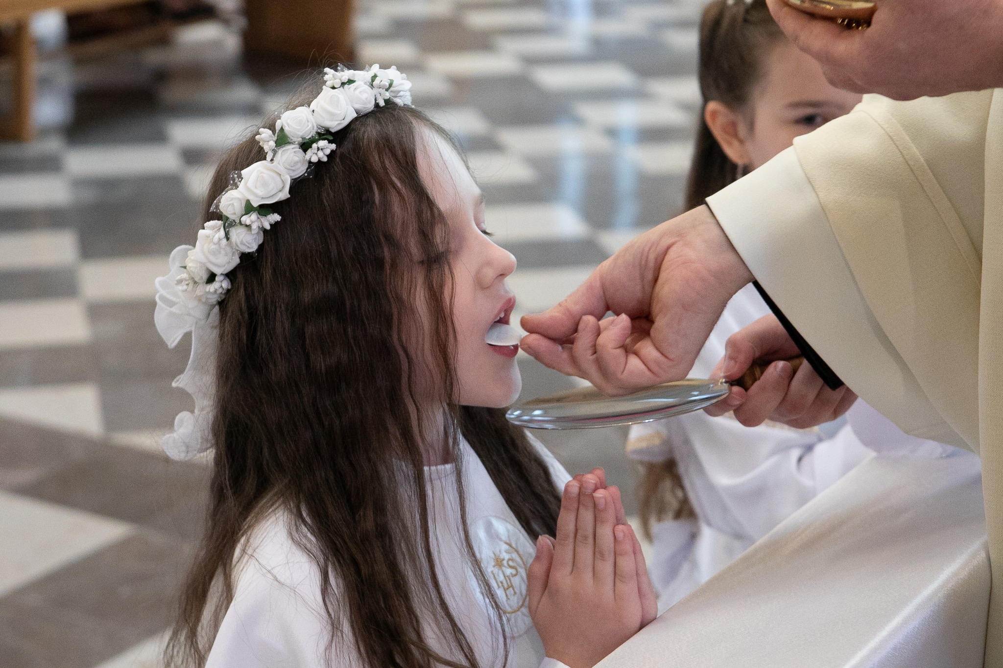 Dziewczynka w białej sukience i wianku przyjmuje komunię świętą. Ujęcie z profilu, widoczne złożone dłonie i srebrna patena. W tle posadzka w szachownicę i inna osoba.