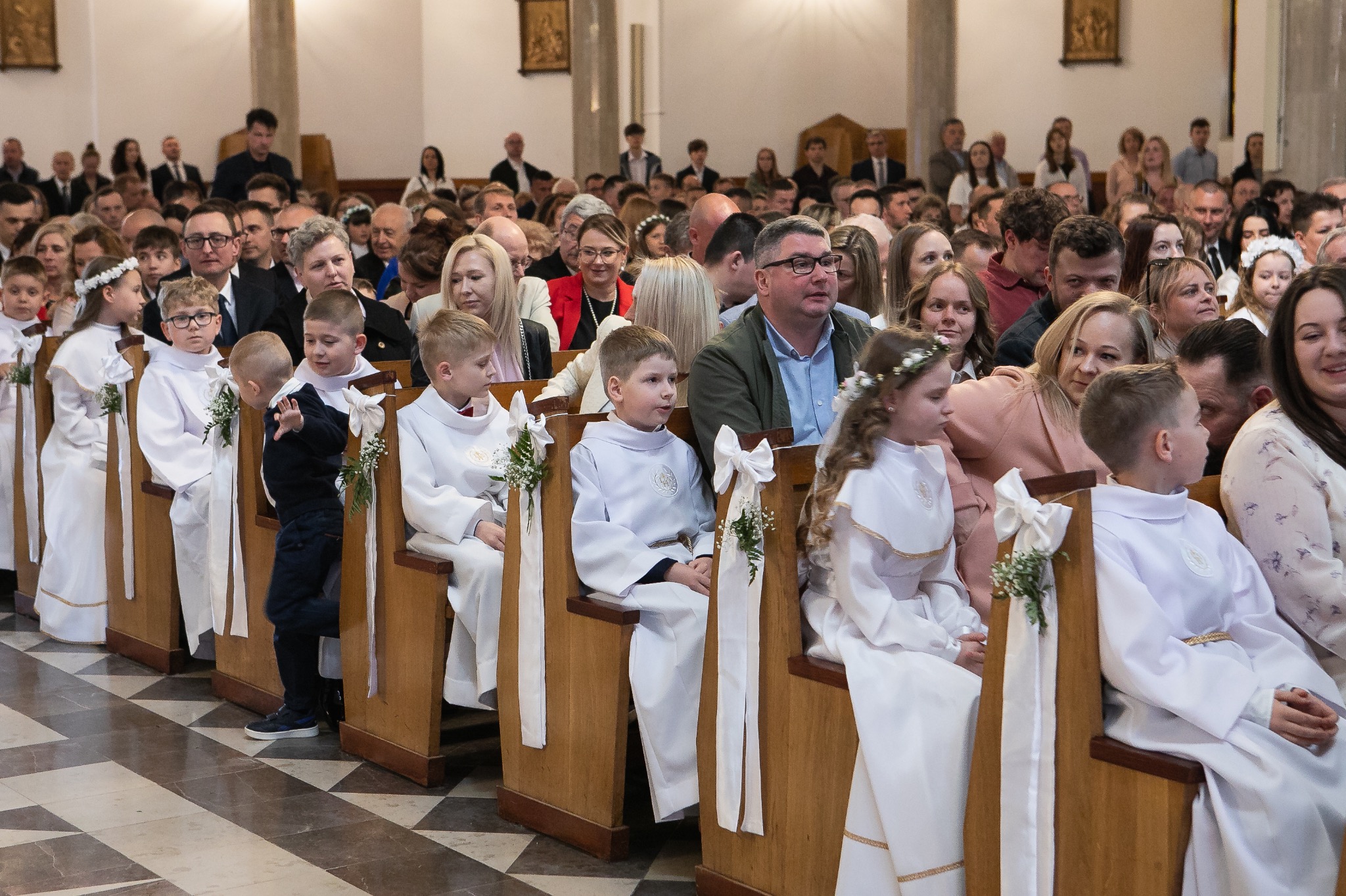 Uczestnicy Pierwszej Komunii Świętej siedzą w ławkach kościoła, w tle zgromadzeni goście. Dzieci ubrane w białe alby, ławki ozdobione białymi wstążkami.