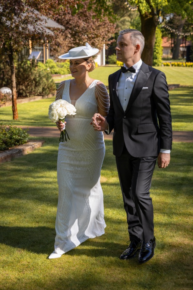 Elegancka para młoda spaceruje po zielonym trawniku w słoneczny dzień. Panna młoda w kapeluszu trzyma bukiet białych róż. Pan młody w klasycznym smokingu.