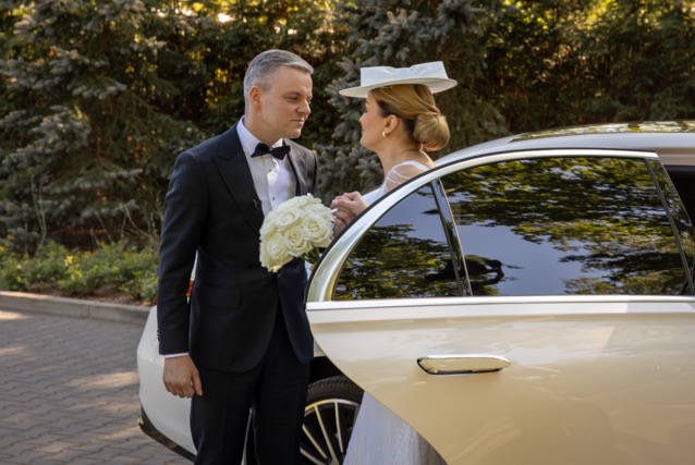 Elegancka para młoda: Pan młody w smokingu z bukietem białych róż spogląda na Pannę Młodą w kapeluszu, wychodzącą z limuzyny w słoneczny dzień ślubu.