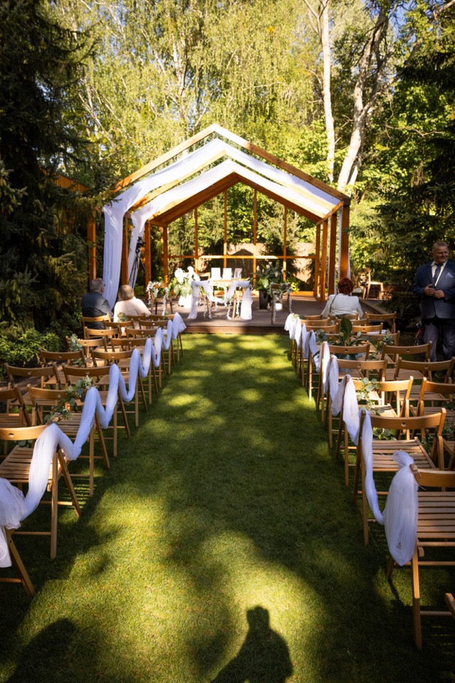 Uroczystość ślubna w plenerze: drewniana altana, krzesła z białymi dekoracjami, zielony trawnik i goście. Cień fotografa w dolnej części kadru.