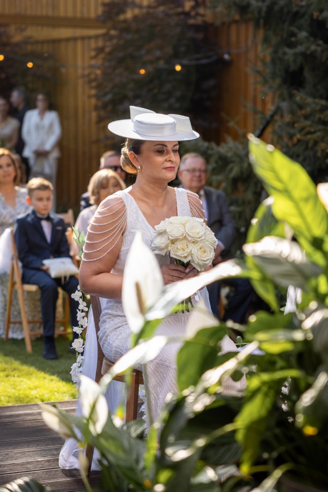 Panna młoda w białej sukni i kapeluszu trzyma bukiet białych róż podczas ceremonii ślubnej w plenerze. W tle goście i zielona roślinność.