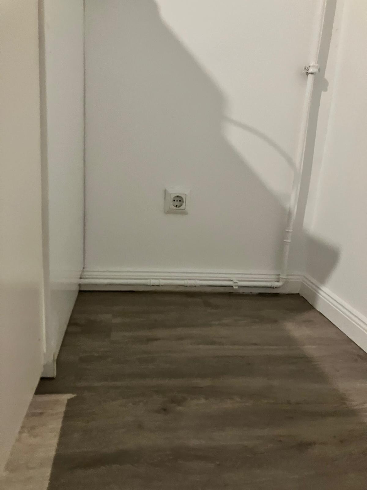 Wnętrze pomieszczenia po remoncie: ciemne panele podłogowe, białe ściany z gniazdkiem elektrycznym i rurami grzewczymi przy podłodze.