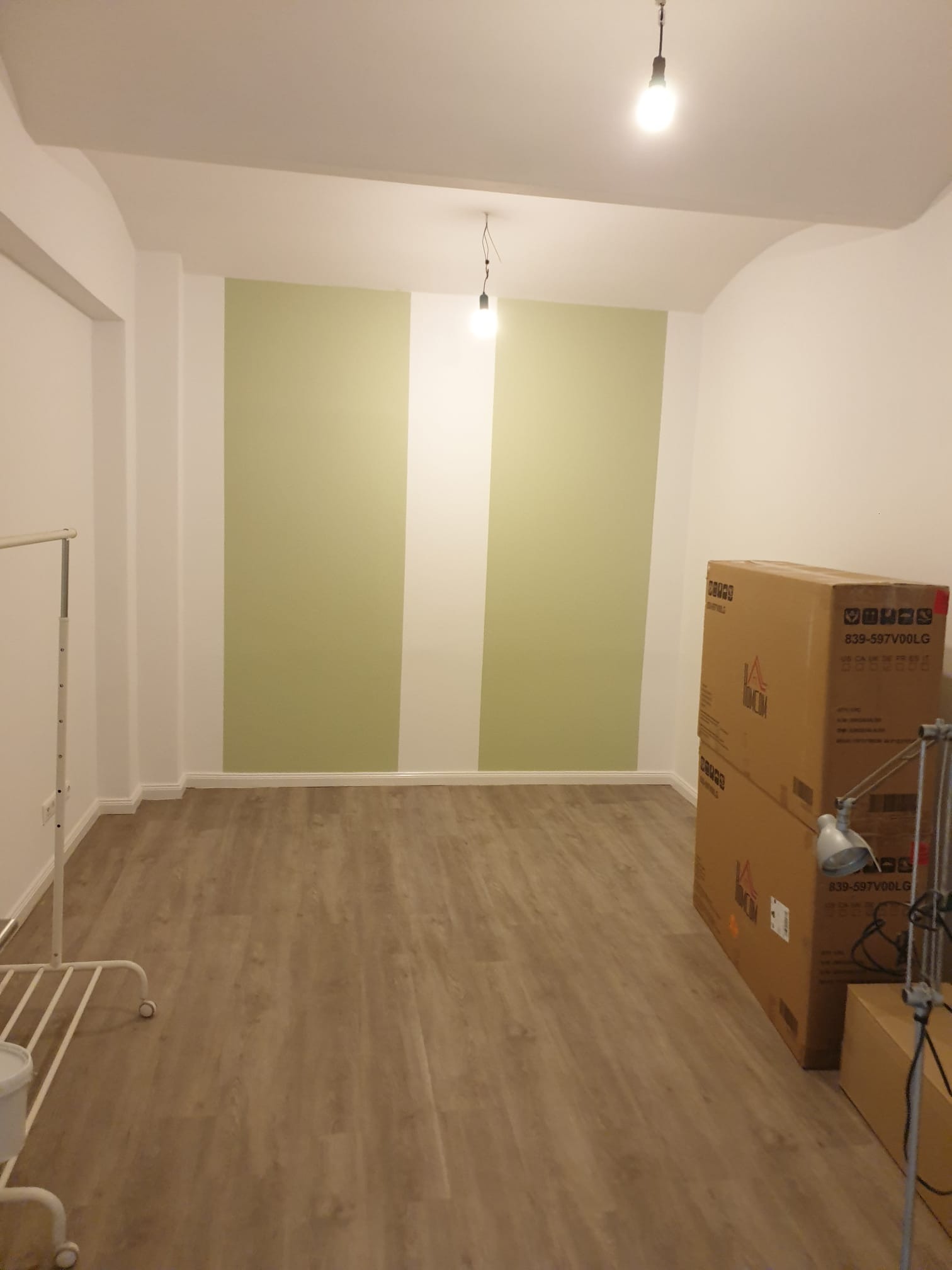 Pomalowane ściany na biało i zielono w nowym pokoju z drewnianą podłogą. Kartony z lampą stoją w rogu. W tle widoczne żarówki.