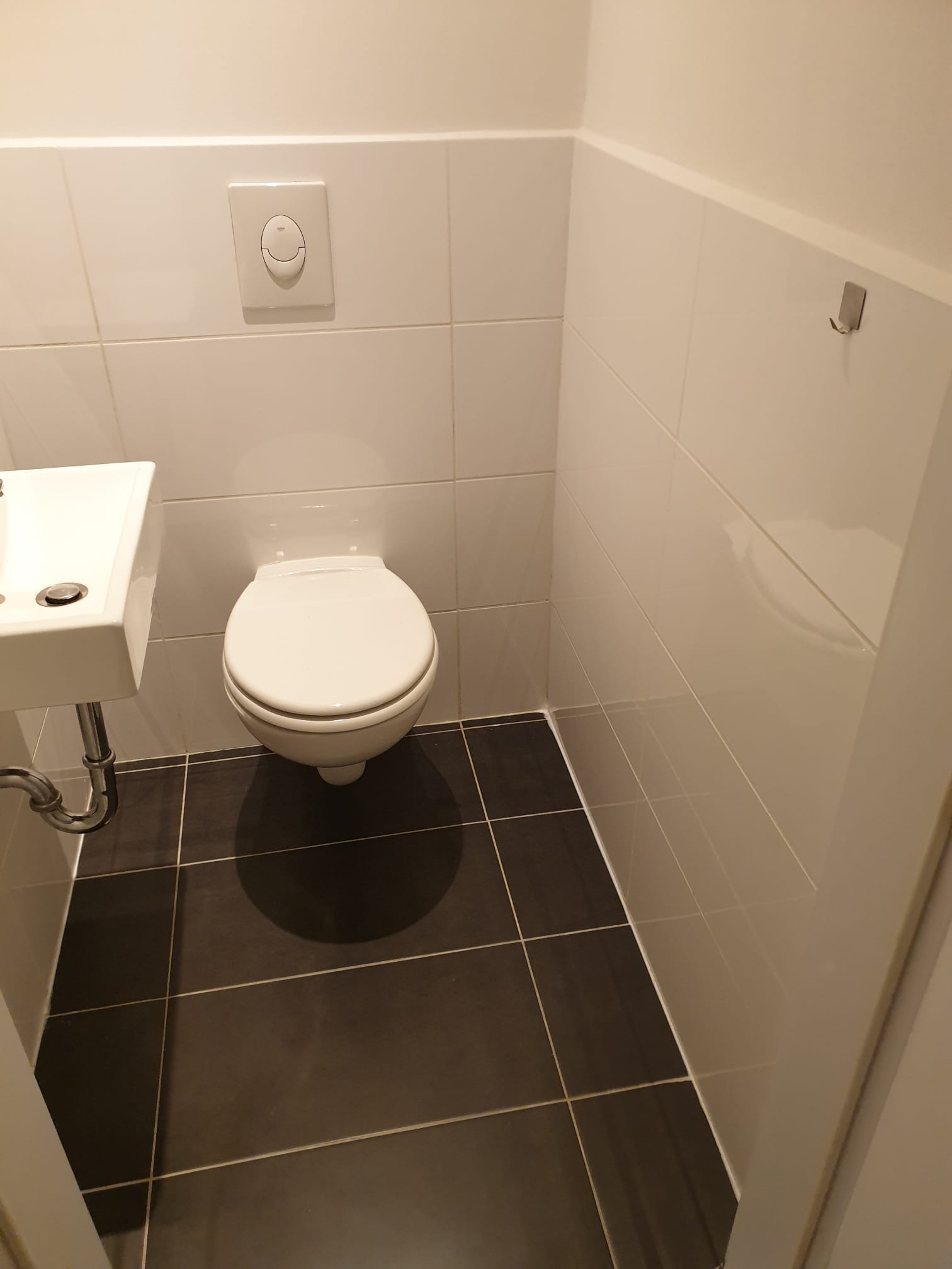 Wnętrze toalety po remoncie: białe płytki ścienne, ciemnobrązowe płytki podłogowe, wisząca umywalka i toaleta. Minimalistyczny design z chromowanymi elementami.