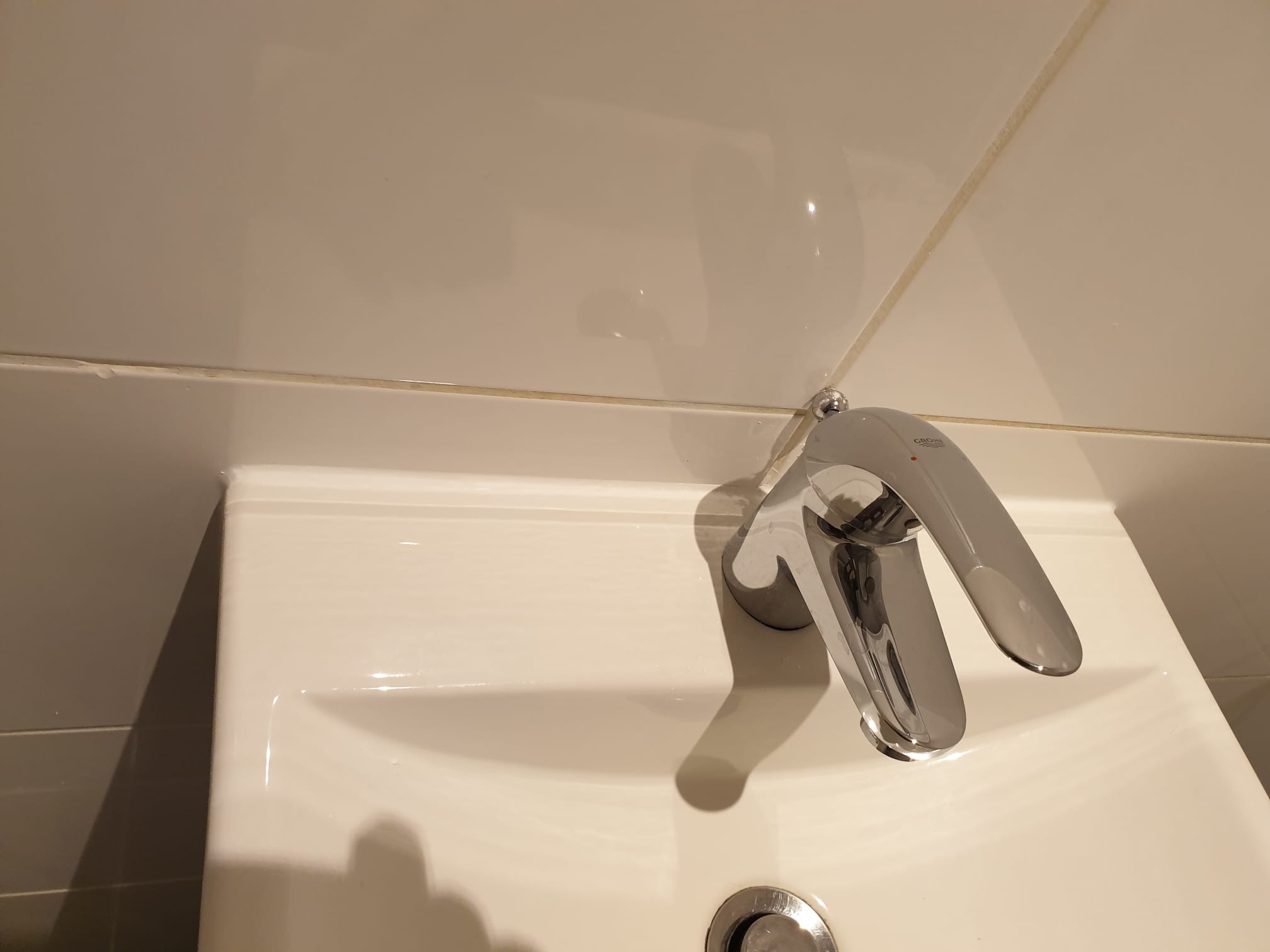 Biała umywalka z chromowaną baterią Grohe, widok z bliska na fugę silikonową między płytkami. Minimalistyczna łazienka po remoncie, neutralne oświetlenie.