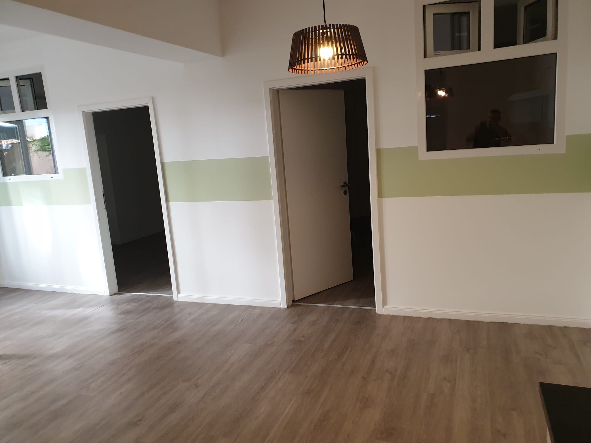 Jasne wnętrze po remoncie: panele podłogowe, białe ściany z zielonym pasem, dwoje drzwi i lampa z drewnianych listewek.