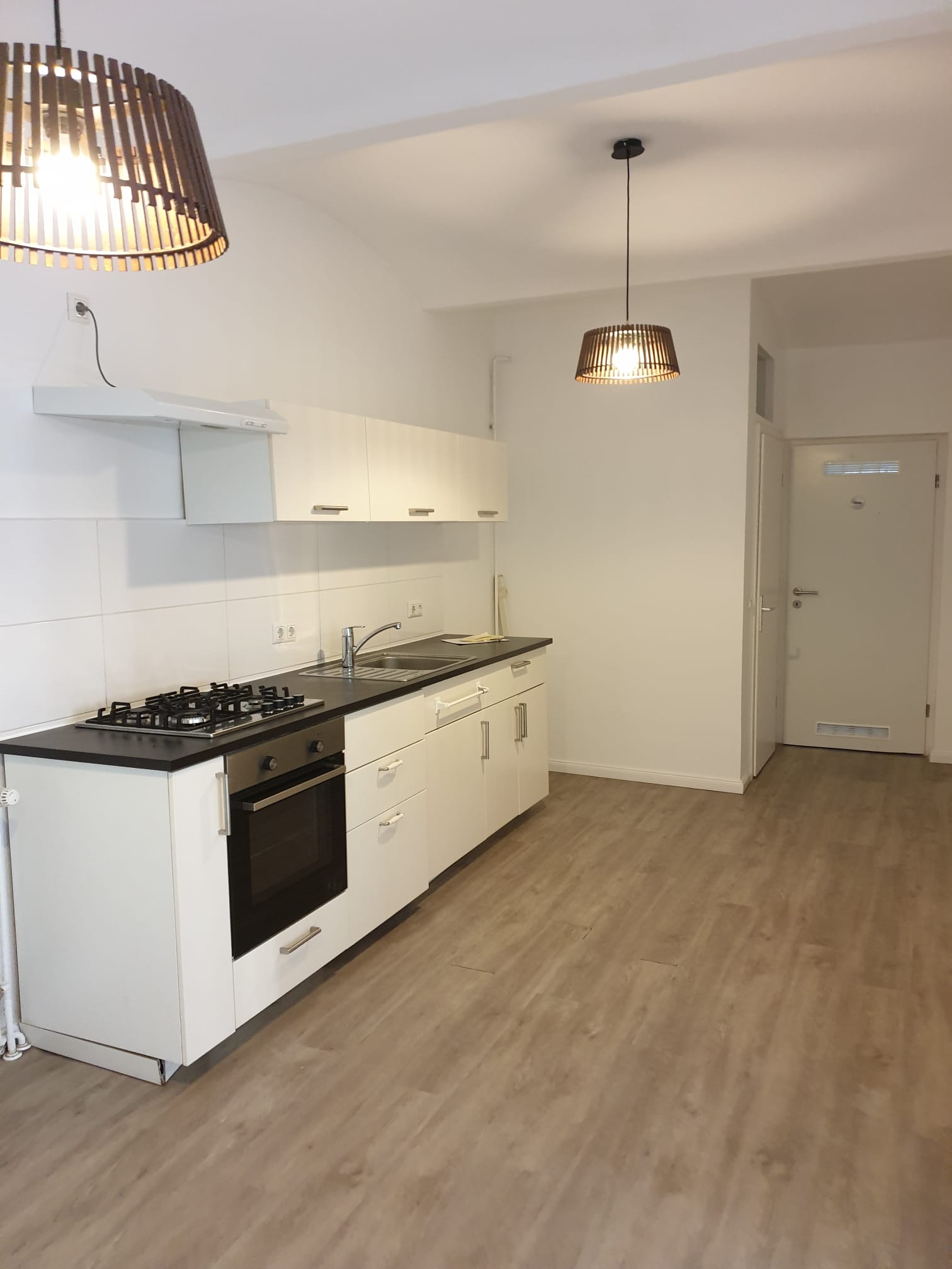 Jasna kuchnia z białymi szafkami, czarnym blatem i drewnianą podłogą. Dwa ażurowe abażury dodają wnętrzu ciepła. Widok na drzwi i część przedpokoju.