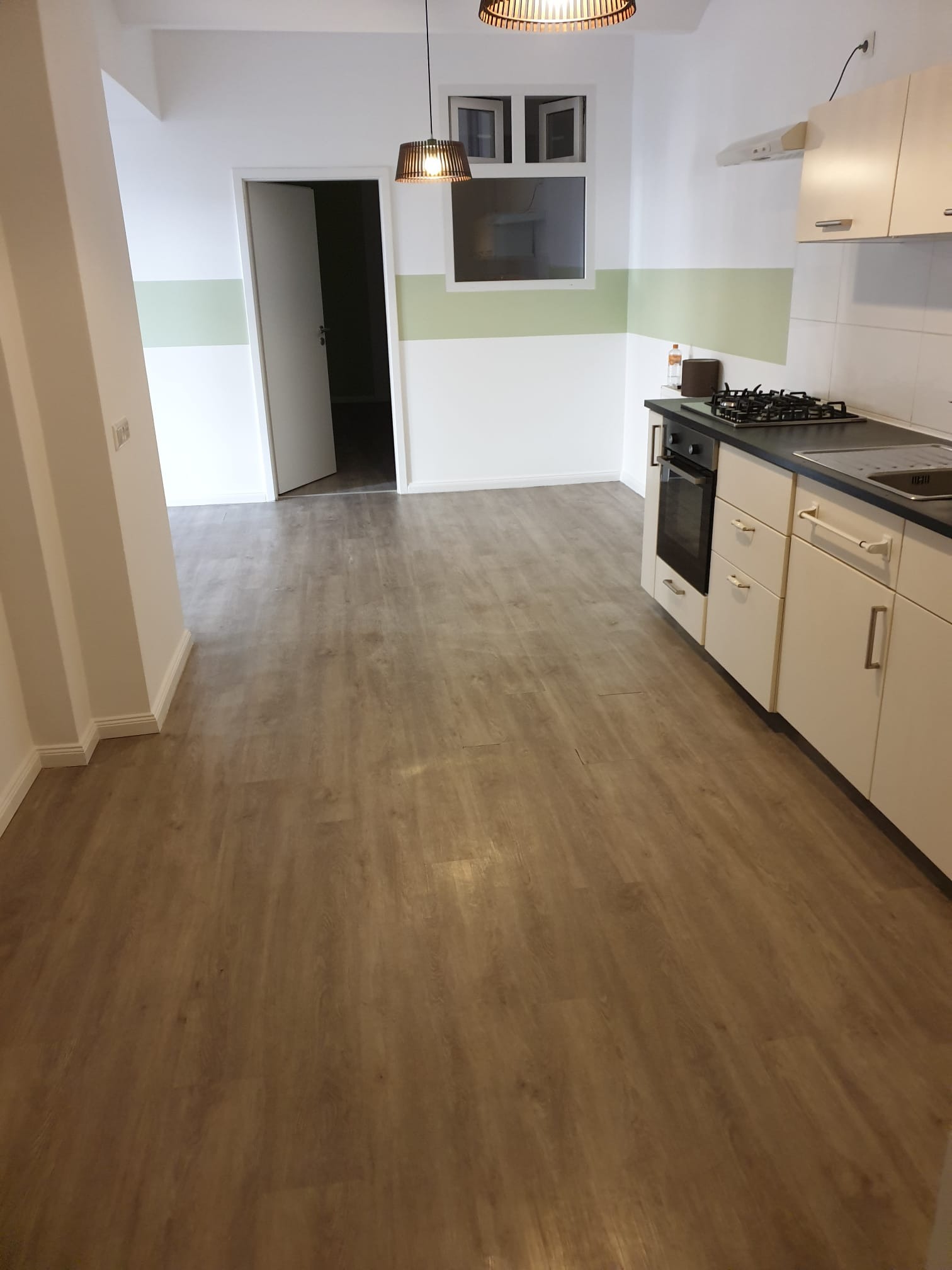 Jasna kuchnia po remoncie z panelami podłogowymi w odcieniach brązu, białe ściany z zielonym pasem i kuchenka w zabudowie. Widok na otwarte drzwi.