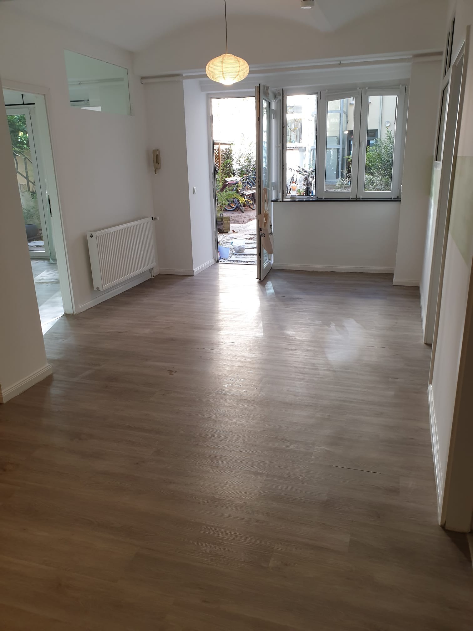 Jasne panele podłogowe w pustym pokoju z białymi ścianami, grzejnikiem i oknem balkonowym wychodzącym na zewnątrz. Widoczne detale wykończenia.