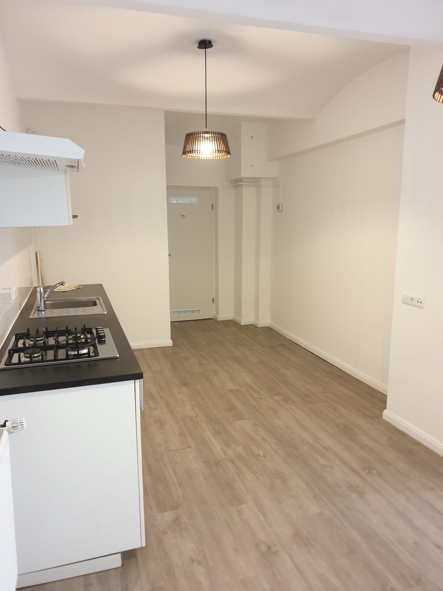 Jasna kuchnia po remoncie. Nowe panele podłogowe, białe szafki z czarnym blatem i stalowa płyta grzewcza. Lampa wisząca z drewnianymi elementami.