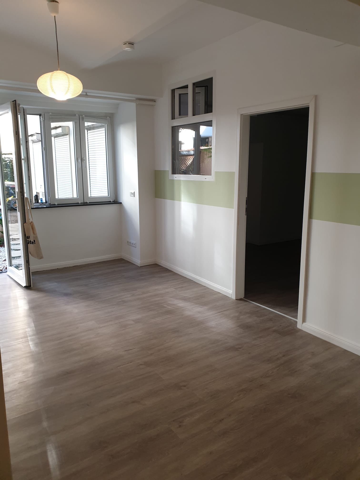 Jasne wnętrze po remoncie: panele podłogowe, białe ściany z zielonym pasem, okna z żaluzjami i otwarte drzwi. Lampa sufitowa w stylu skandynawskim.