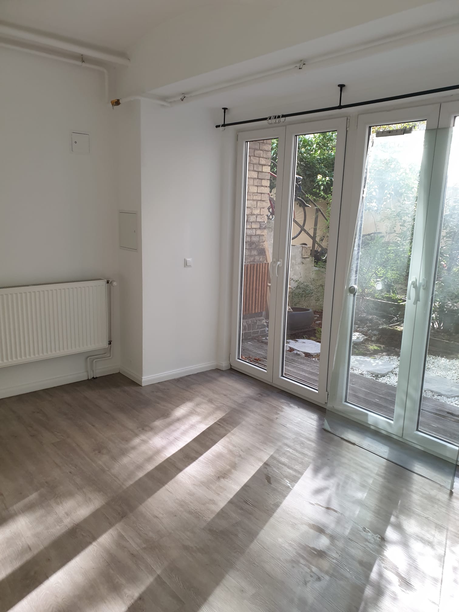 Jasne, przestronne wnętrze po remoncie: panele podłogowe, białe ściany, grzejnik i duże okno balkonowe z widokiem na ogród. Promienie słońca na podłodze.