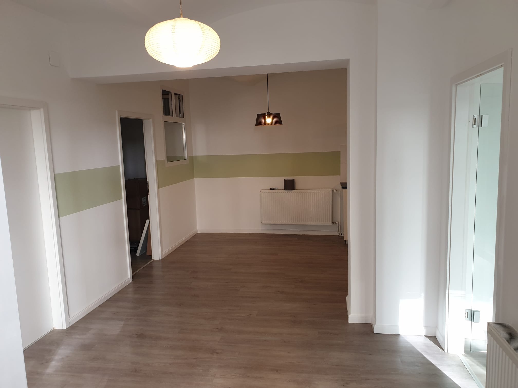 Wykończone wnętrze mieszkania po remoncie: panele podłogowe, pomalowane ściany z poziomym pasem koloru, drzwi szklane i białe, lampy sufitowe.