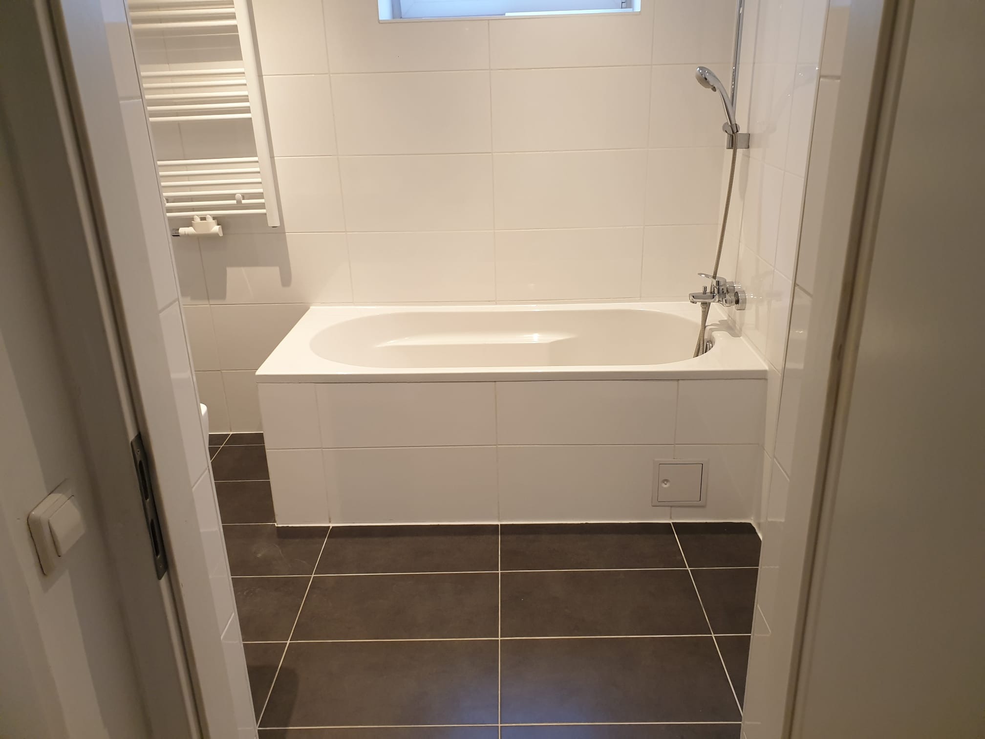 Wykończona łazienka z białą wanną, grzejnikiem drabinkowym i brązowymi płytkami podłogowymi. Ujęcie zza uchylonych drzwi.