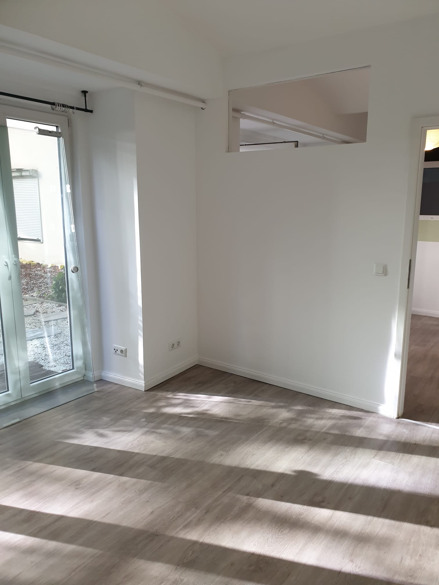 Jasne panele podłogowe w pustym pokoju z białymi ścianami i dużym oknem balkonowym. Widoczne gniazdka elektryczne i otwarte przejście do innego pomieszczenia.