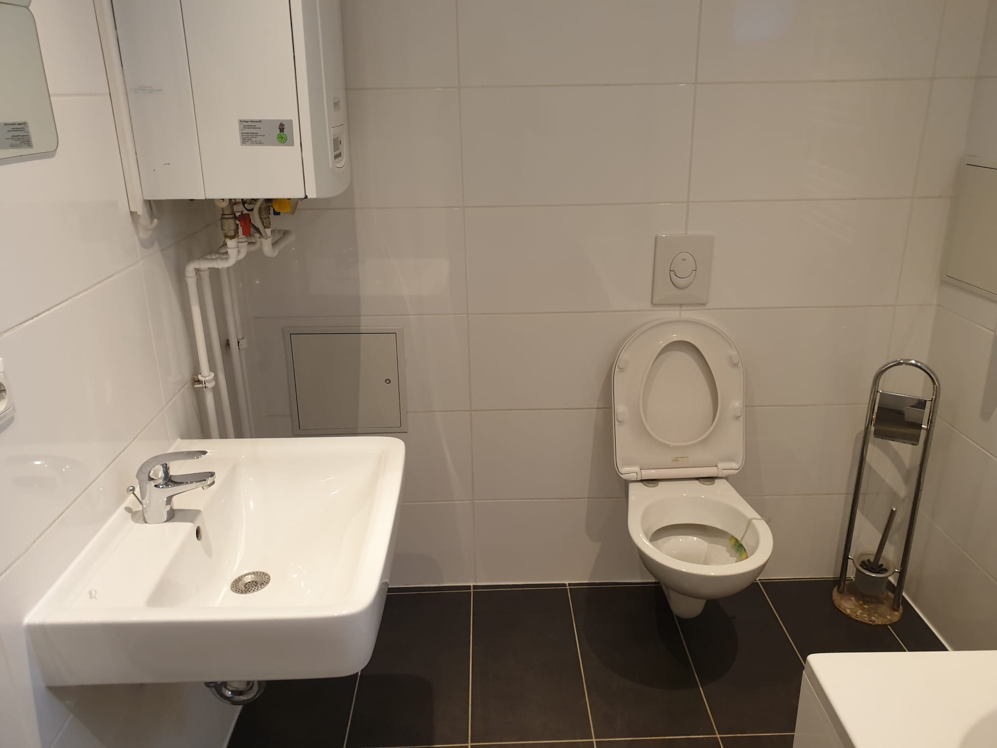 Wykończona łazienka: biała umywalka z chromowaną baterią, toaleta z otwartą klapą, grzejnik i ciemna podłoga z fugami. Minimalistyczny design.