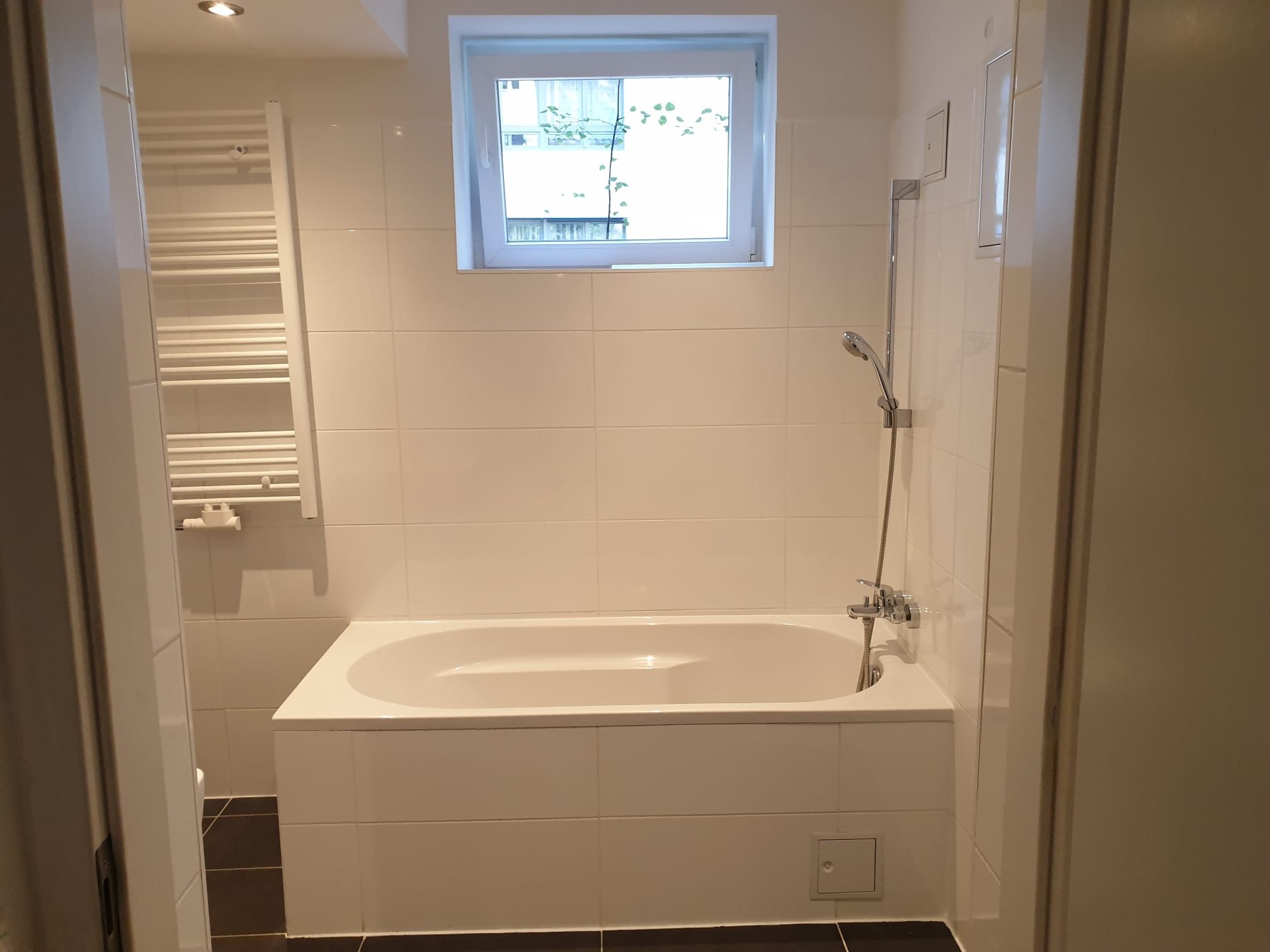 Jasna łazienka po remoncie: białe płytki, wanna obudowana płytkami, okno z widokiem na zieleń, grzejnik drabinkowy. Minimalistyczny design.