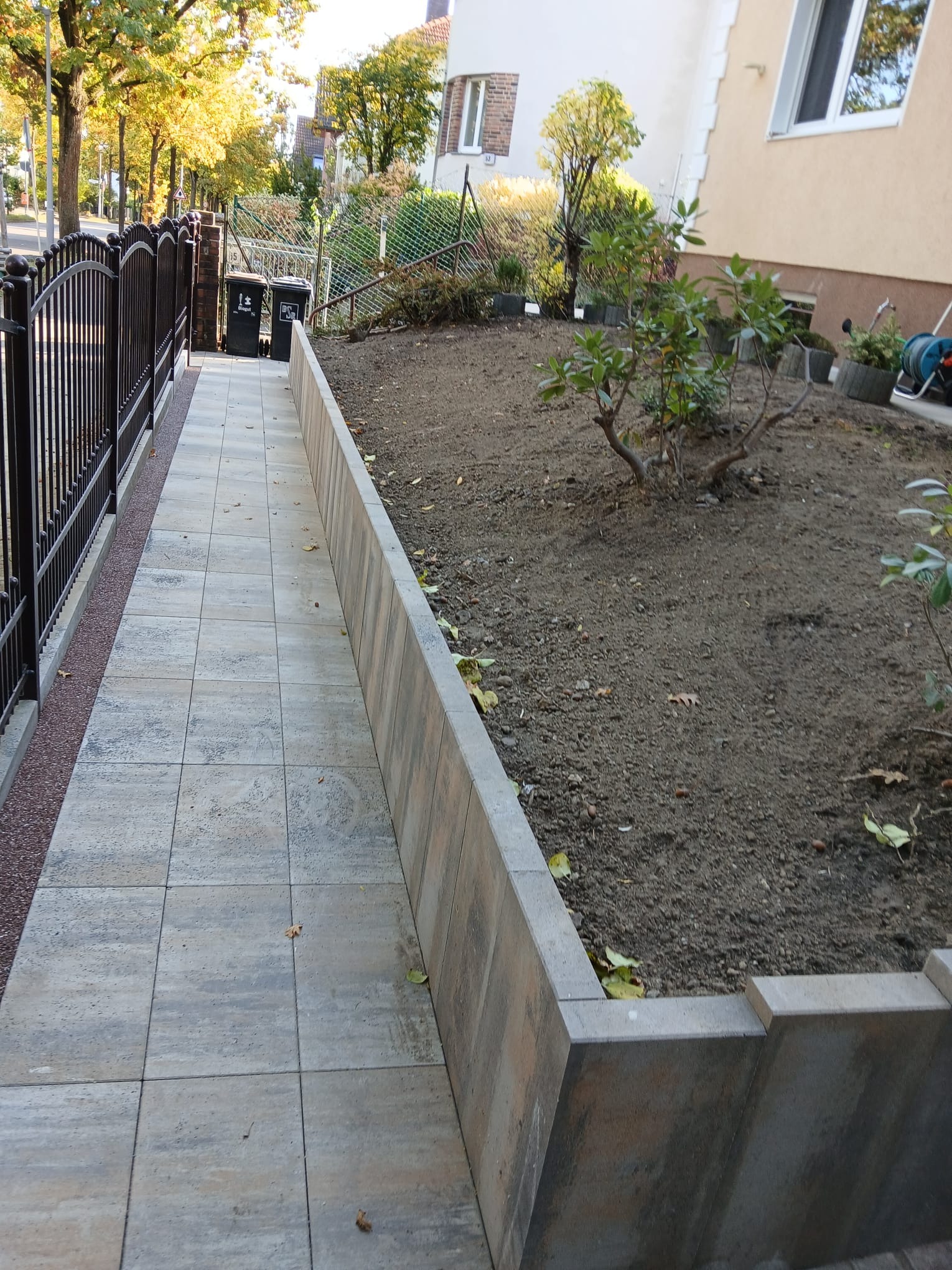 Ułożona kostka brukowa tworząca ścieżkę wzdłuż posesji, z widocznym ogrodzeniem i rabatą ziemi. Regularny wzór kostki, stonowana kolorystyka, jesienne liście.