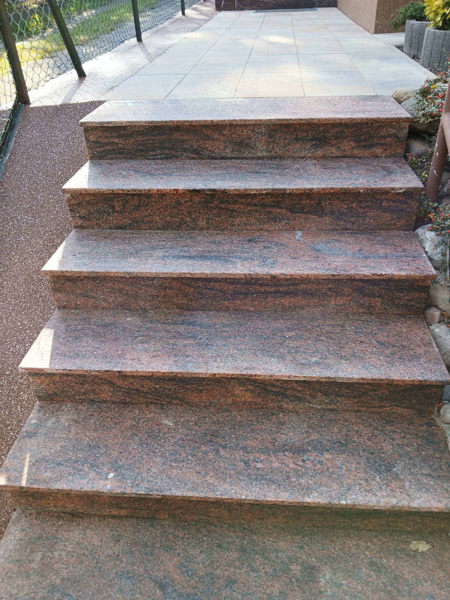 Granitowe schody zewnętrzne z widoczną fakturą kamienia, prowadzące do wyżej położonego chodnika z jasnych płyt. Po bokach widoczne elementy zieleni i ogrodzenia.