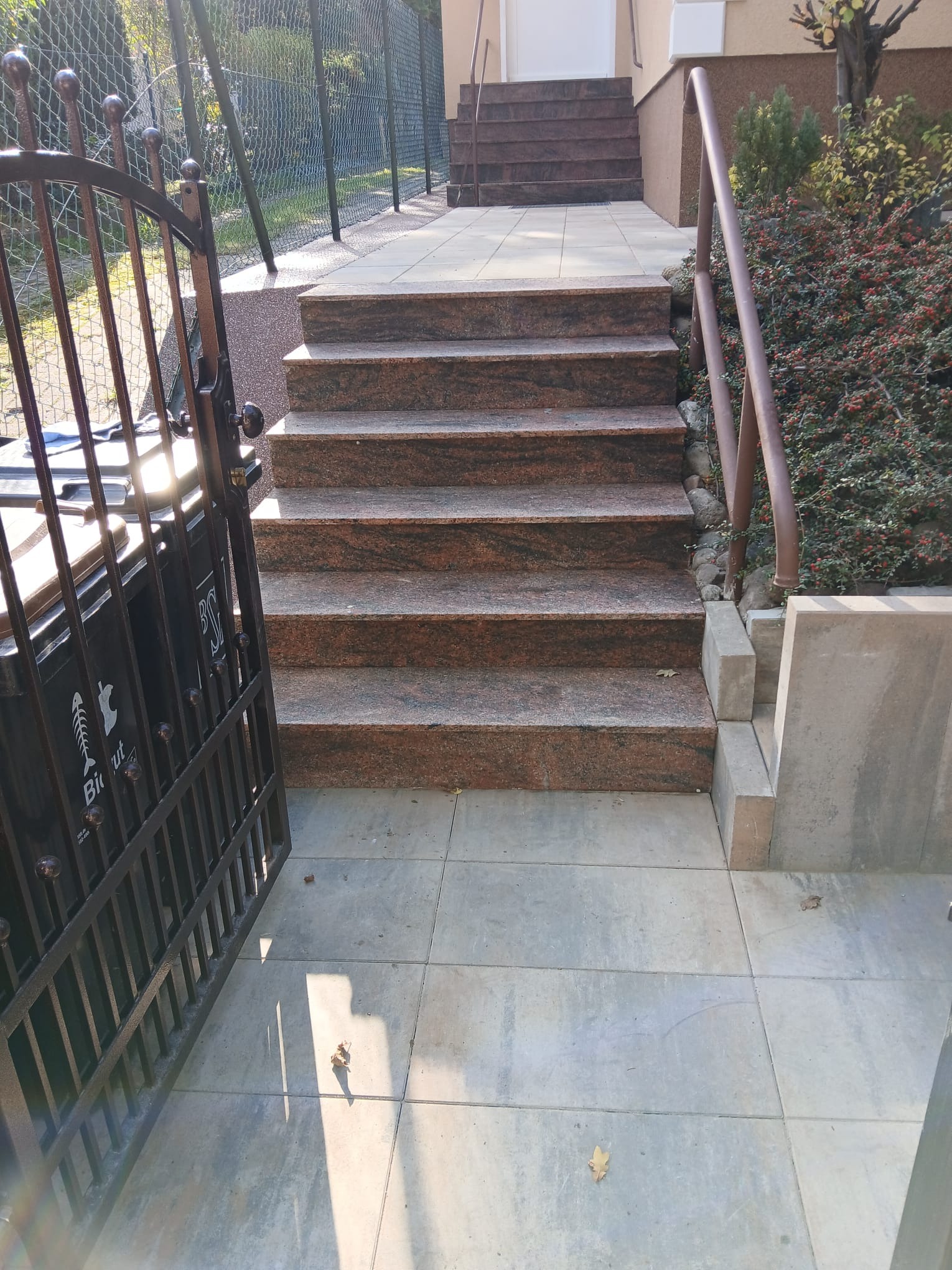 Zewnętrzne schody z czerwonego granitu, prowadzące do domu. Widoczne płytki na podłożu, metalowa brama i fragment zieleni. Słoneczny dzień.