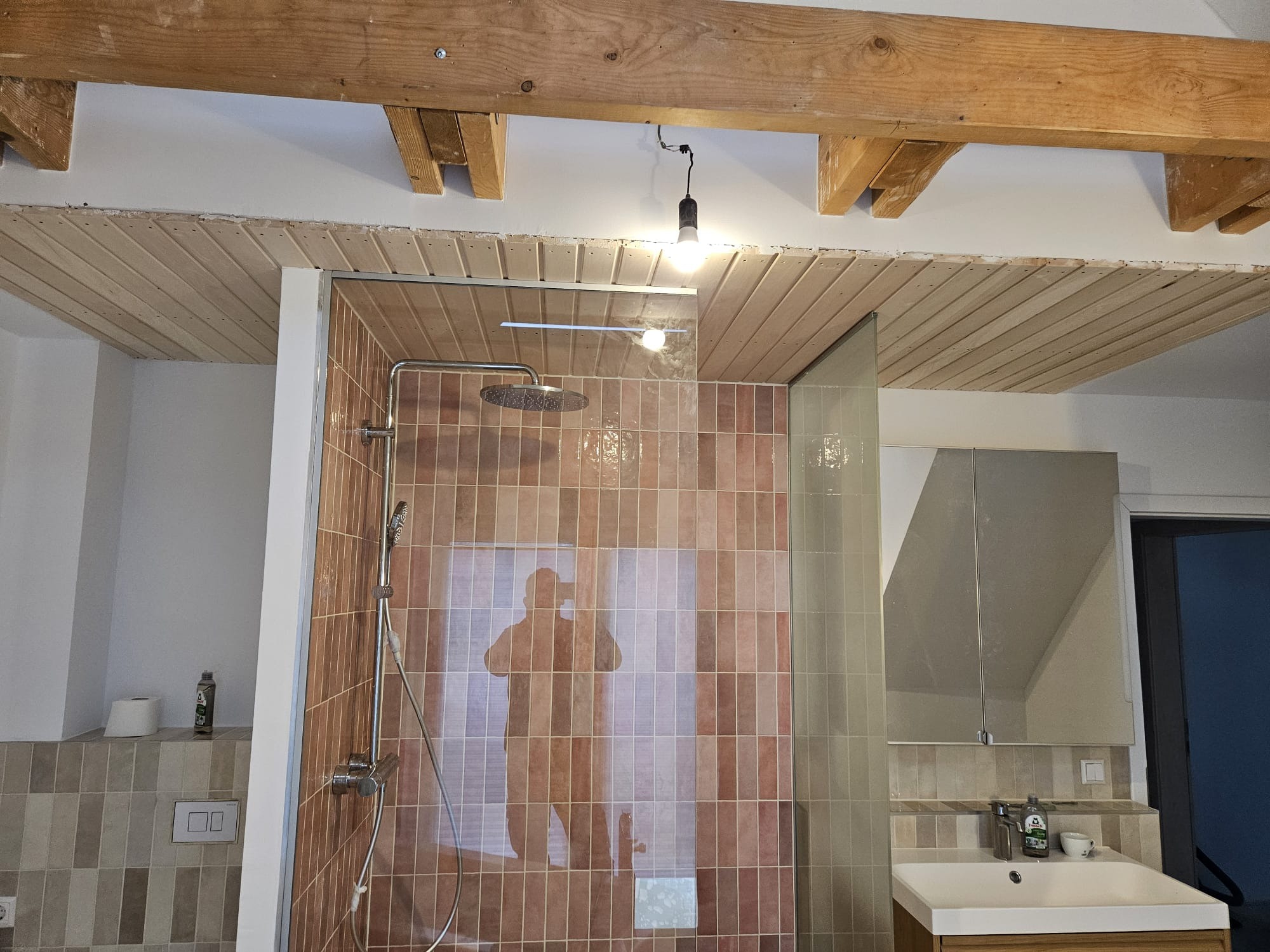 Wykończona kabina prysznicowa z brązowymi płytkami, drewnianym sufitem i belkami. Widoczne odbicie osoby robiącej zdjęcie. Umywalka z lustrem w tle.