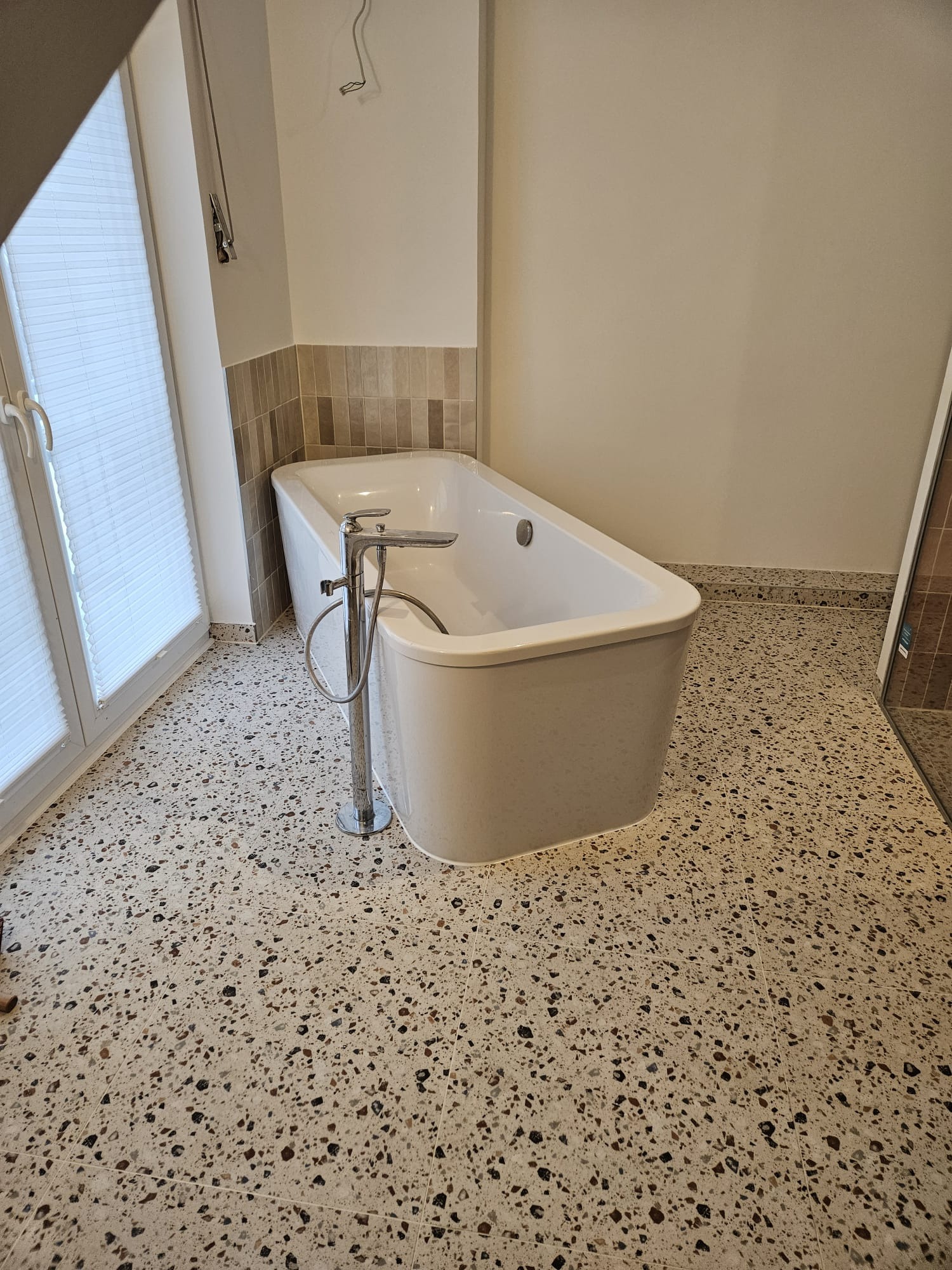 Nowoczesna, wolnostojąca wanna z chromowaną armaturą na tle beżowej ściany z dekoracyjnym pasem płytek i podłogi lastryko. Widok na okno z żaluzjami.