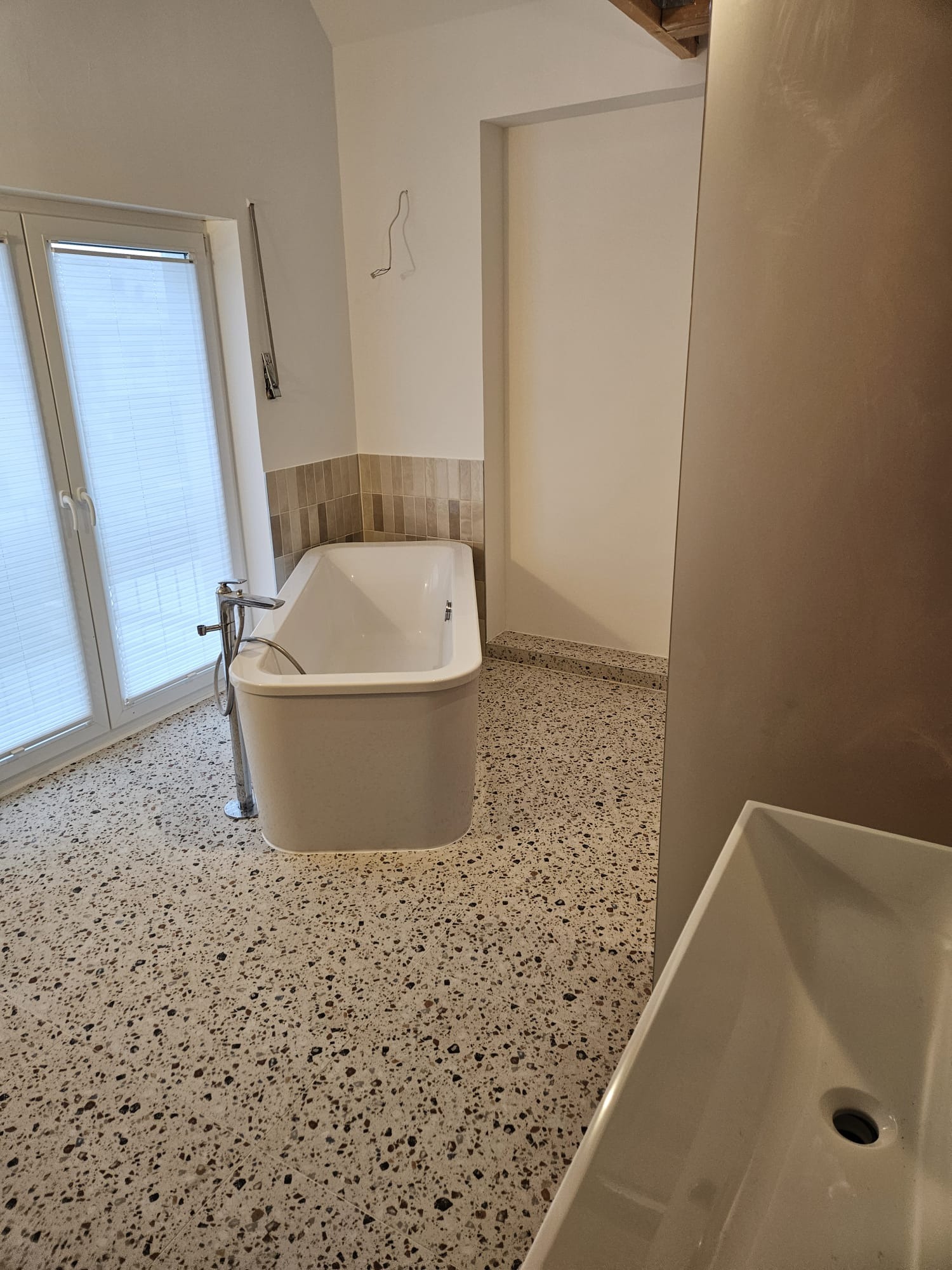 Nowoczesna łazienka z wolnostojącą wanną, mozaikową podłogą i minimalistyczną armaturą. Widoczne okno z żaluzjami i fragment umywalki.