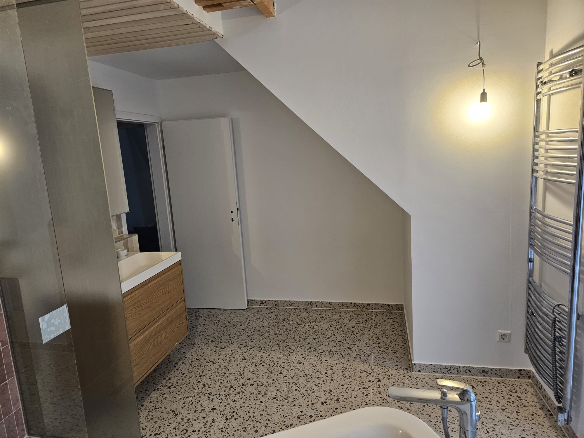 Nowoczesna łazienka po remoncie: terazzo na podłodze, drewniana szafka pod umywalką, grzejnik drabinkowy i minimalistyczna lampa wisząca. Widok na otwarte drzwi.