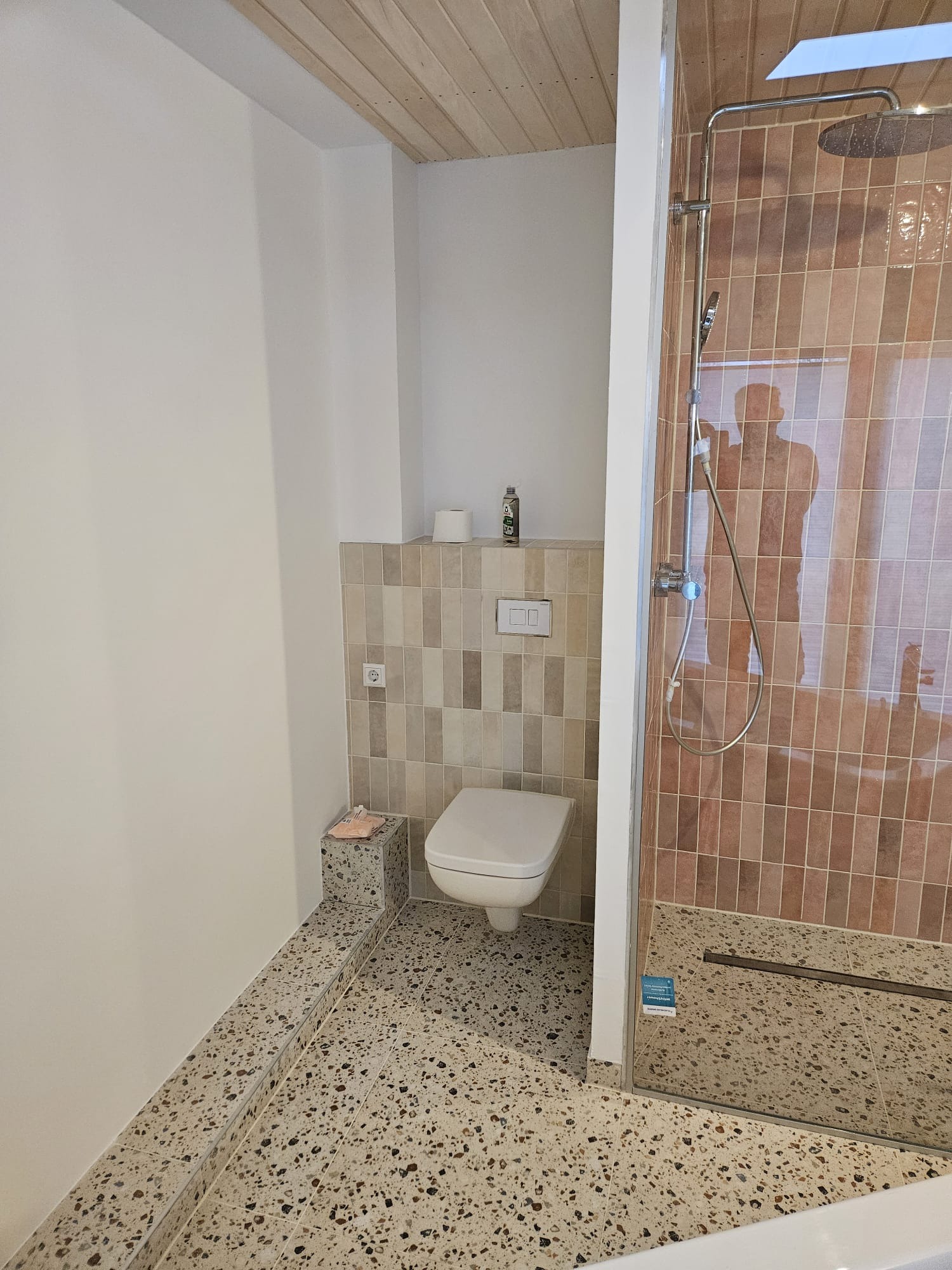 Nowoczesna łazienka po remoncie: beżowe płytki w kabinie prysznicowej, mozaika na podłodze i ścianie za toaletą. Drewniana boazeria na suficie.