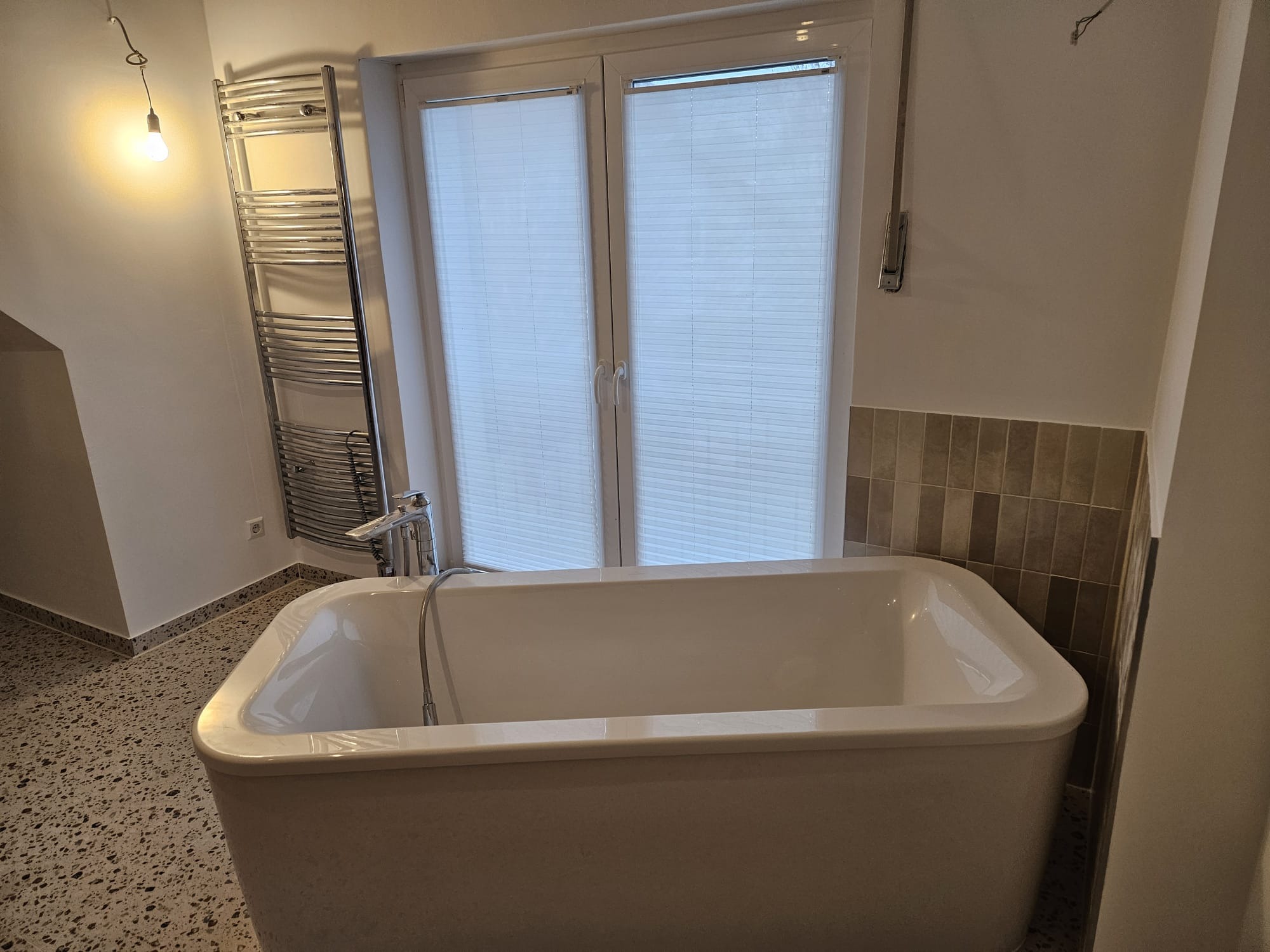 Nowoczesna, wolnostojąca wanna z chromowaną baterią i grzejnikiem drabinkowym w wykończonej łazience z oknem i żaluzjami plisowanymi. Terrazzo na podłodze.