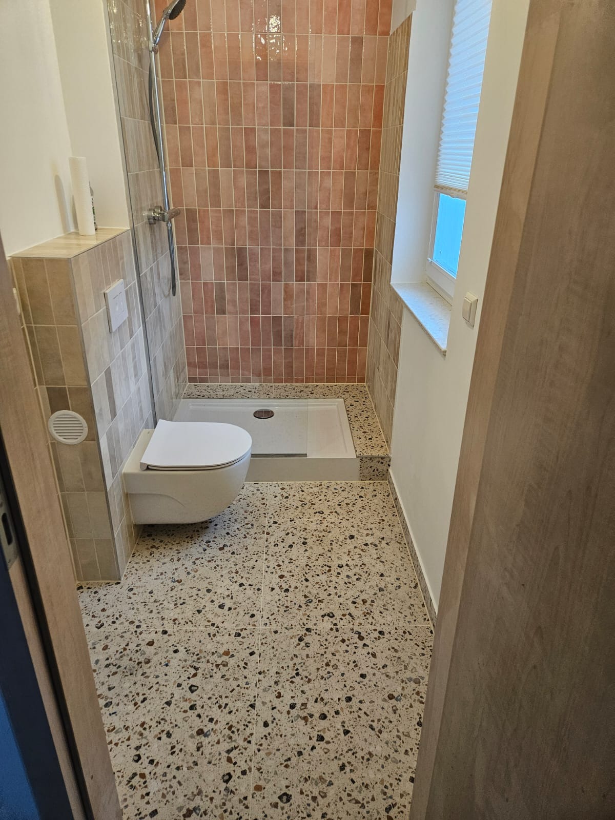 Wykończona łazienka z kabiną prysznicową, toaletą podwieszaną i terakotową mozaiką na ścianach. Widok zza uchylonych drzwi, okno z żaluzjami w tle.