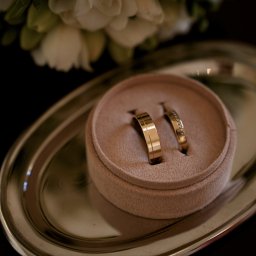 IOT DEVHOUSE SPÓŁKA Z OGRANICZONĄ ODPOWIEDZIALNOŚCIĄ - Dwa złote pierścionki ślubne, jeden z brylantami, leżące w beżowym pudełku na srebrnej tacy. W tle rozmyte białe kwiaty. Delikatne światło.