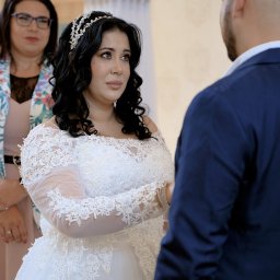 IOT DEVHOUSE SPÓŁKA Z OGRANICZONĄ ODPOWIEDZIALNOŚCIĄ - Panna młoda w koronkowej sukni spogląda na pana młodego podczas ceremonii ślubnej. W tle kobieta w kwiecistej marynarce. Ujęcie z bliska, skupione na emocjach.