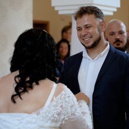 IOT DEVHOUSE SPÓŁKA Z OGRANICZONĄ ODPOWIEDZIALNOŚCIĄ - Uśmiechnięty pan młody w granatowym garniturze spogląda na pannę młodą w białej sukni z koronkowymi zdobieniami. W tle goście weselni i kolumna.