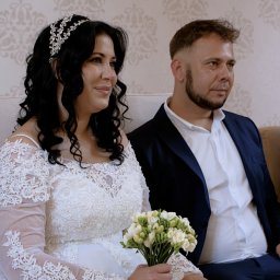 IOT DEVHOUSE SPÓŁKA Z OGRANICZONĄ ODPOWIEDZIALNOŚCIĄ - Para młoda siedzi obok siebie: panna młoda w koronkowej sukni trzyma bukiet frezji, obok pan młody w granatowym garniturze i białej koszuli. W tle wzorzysta tapeta.