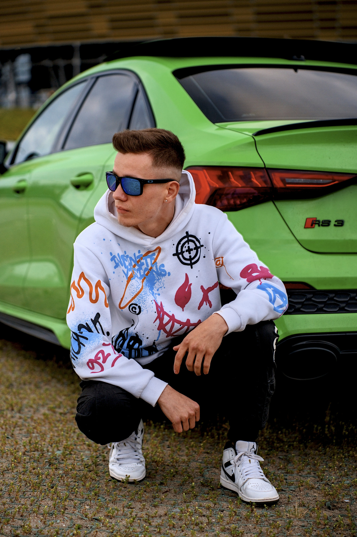 Młody mężczyzna w okularach i bluzie z grafiką, kuca na tle zielonego Audi RS3. Ujęcie z dołu, podkreślające auto i stylizację modela. Dynamiczna poza, miejski styl.