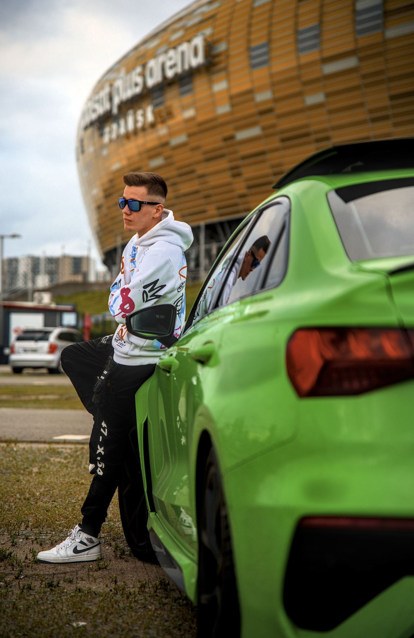Mężczyzna w okularach, oparty o zielony samochód na tle stadionu w Gdańsku. Ubrany w bluzę z nadrukiem i czarne spodnie. Widoczne buty sportowe i saszetka nerka.