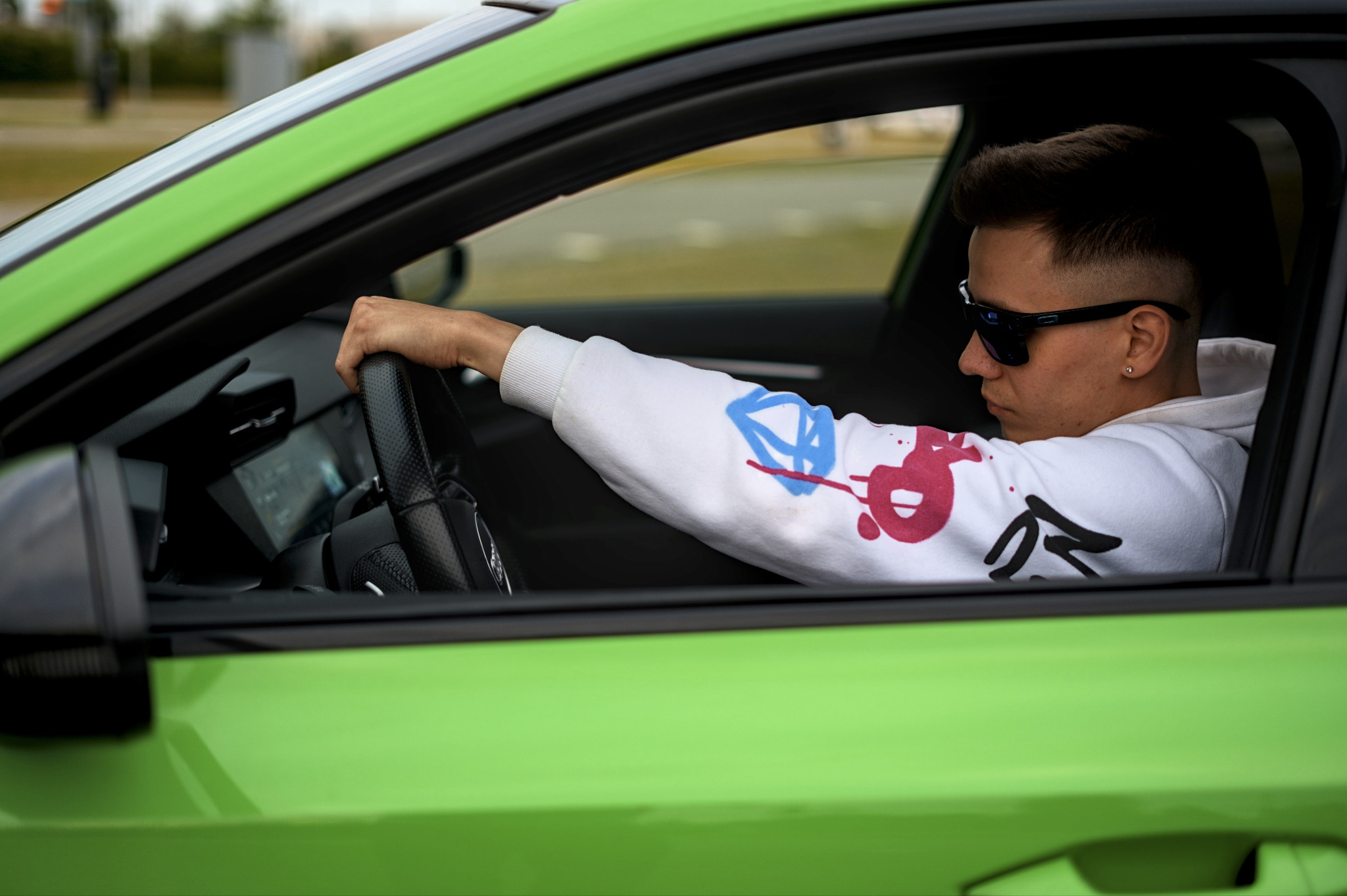 Młody mężczyzna w okularach przeciwsłonecznych prowadzi jaskrawozielony samochód, ubrany w białą bluzę z kolorowymi wzorami. Widok z boku, skupienie na twarzy i dłoni na kierownicy.