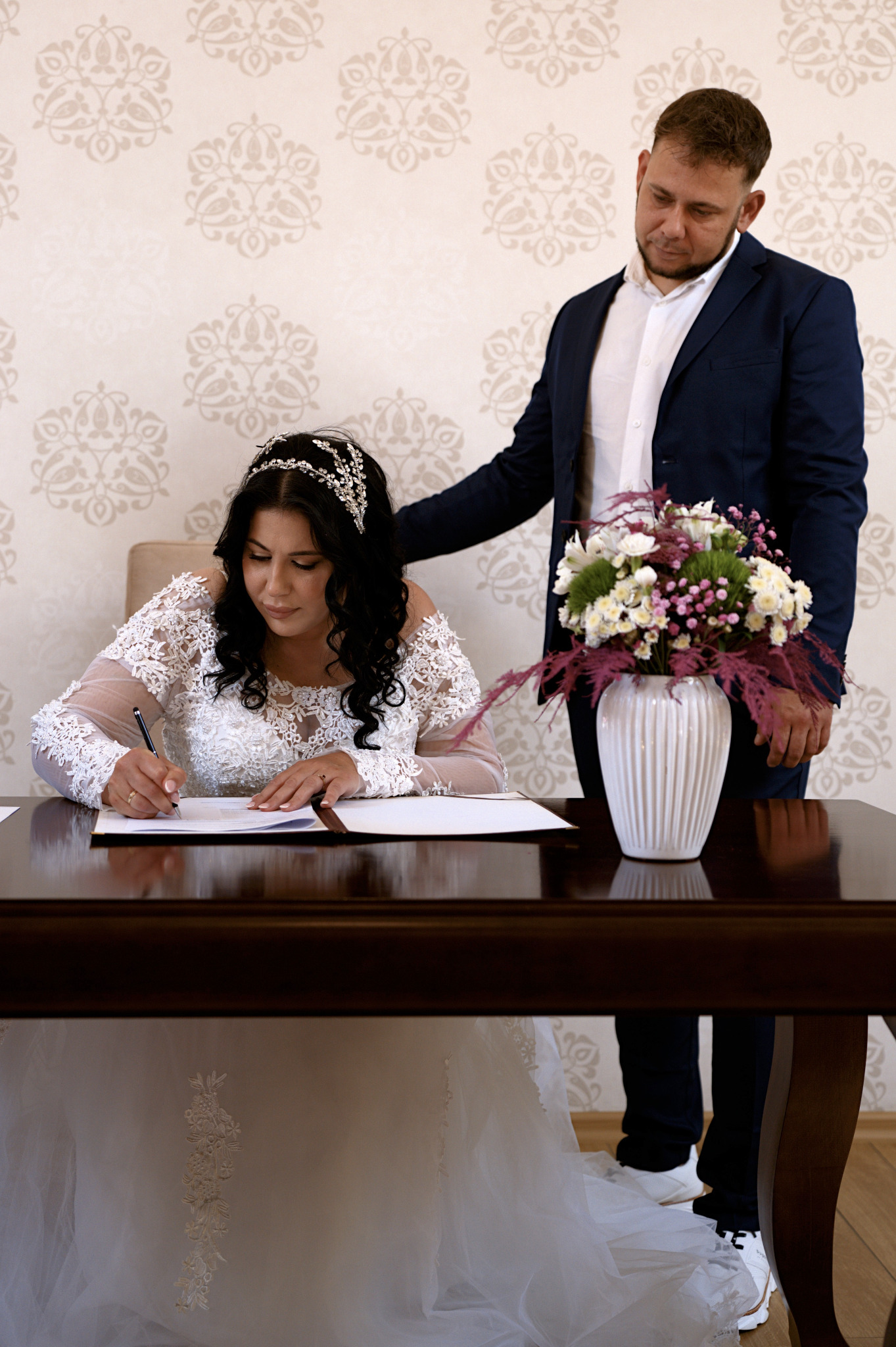 Panna młoda w koronkowej sukni podpisuje dokument, obok stoi pan młody w granatowym garniturze, na stole wazon z kwiatami. Uroczysty moment zawarcia związku małżeńskiego.